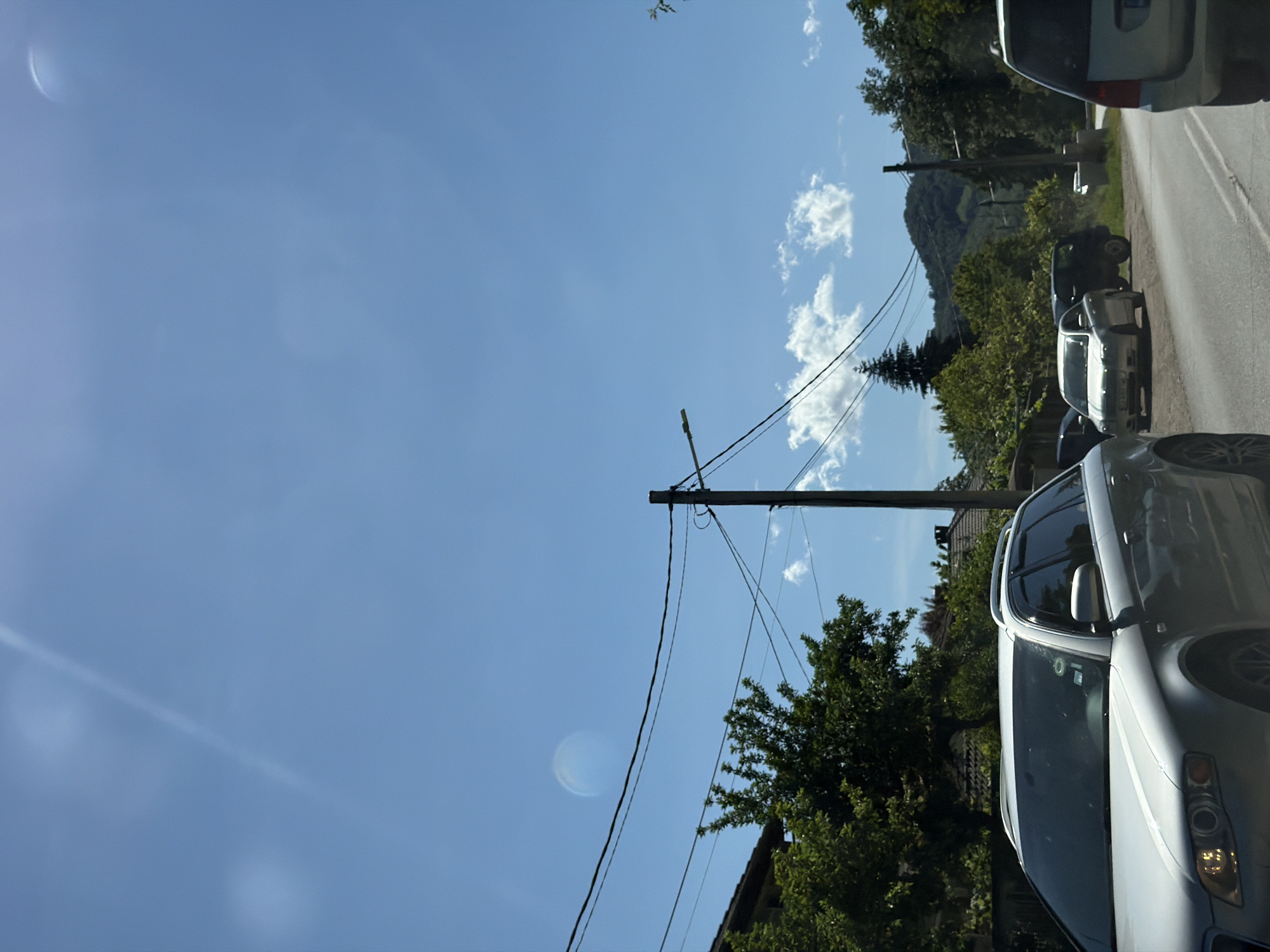 The image captures a view of a street in Beli Osam, Bulgaria, on a sunny day. The sky is a vibrant blue with scattered white clouds. Power lines are visible, strung between utility poles, and a street lamp is mounted on one of them. In the foreground, a silver station wagon is parked. Further down the street, other vehicles are present, including a silver sedan and a dark SUV. Trees and houses line the sides of the road, and in the background, lush green hills rise, indicating a natural, possibly rural or semi-rural setting. The overall impression is one of a typical village scene on a bright, clear day.