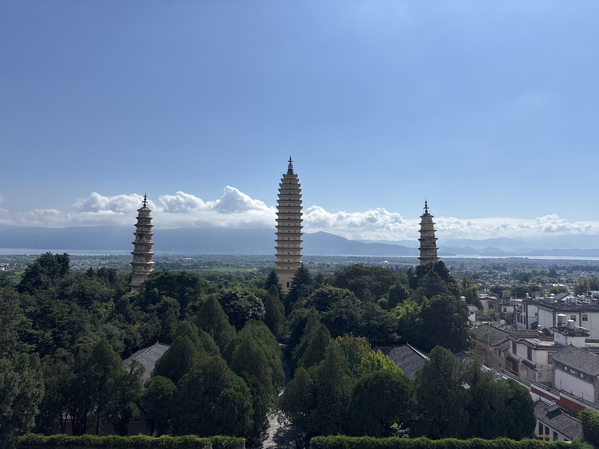 The image captures a panoramic view of the Three Pagodas of Chongsheng Temple, a prominent landmark in Dali City, China. The three ancient pagodas, varying in height and design, stand tall amidst lush green trees and a serene landscape. The pagodas are situated on a hill overlooking the city, with a body of water, likely Erhai Lake, and distant mountains forming the backdrop. The sky is a clear blue, with scattered white clouds, suggesting a pleasant day. The architecture of the pagodas is distinctive, with multiple tiers and intricate detailing. The surrounding vegetation is dense, with a variety of trees and manicured hedges. In the foreground, traditional Chinese architecture with tiled roofs can be seen, characteristic of the region. There are no people visible in the image, emphasizing the tranquility and historical significance of the site. The overall impression is one of natural beauty, cultural heritage, and architectural grandeur.