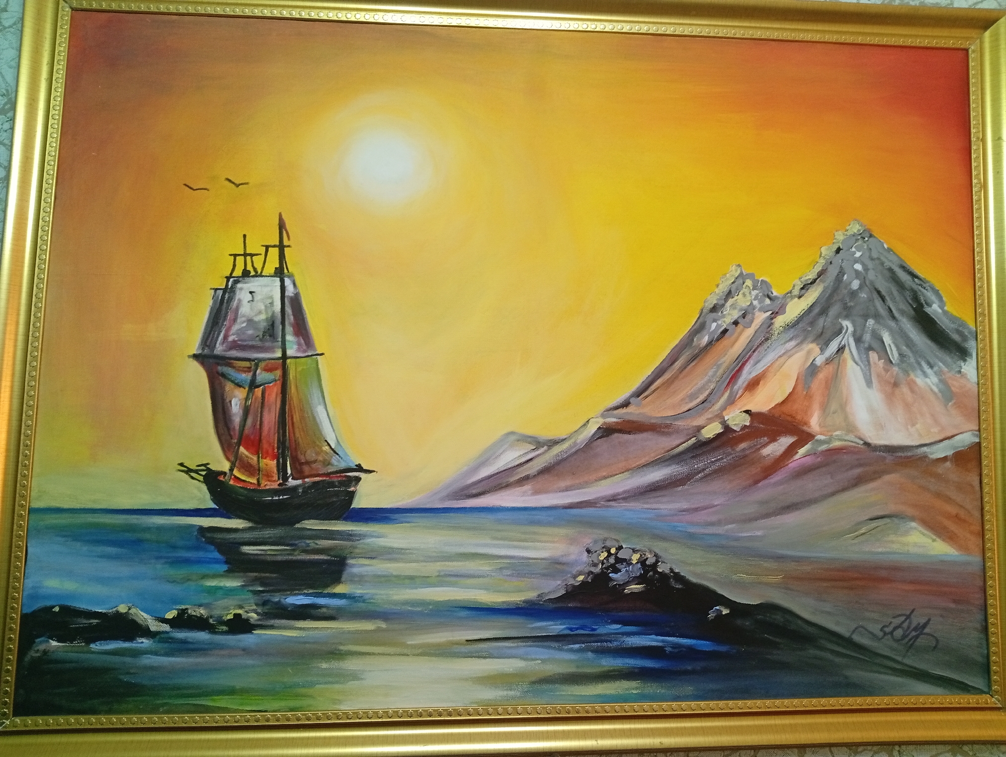 The image is an oil painting framed in gold, depicting a seascape with a sailboat and mountains. The sky is a vibrant gradient of yellow and orange, with a bright, diffused sun in the upper left. Two small black birds are flying in the upper left quadrant. In the center left, a dark sailboat with colorful sails is at sea. Its reflection is visible in the water below. The water is rendered with blues, greens, and whites, suggesting choppy waves. In the right half of the painting, a range of stylized mountains rises from the water. They are painted in shades of gray, purple, orange, and brown, with white accents. In the foreground, a dark, rocky outcrop is visible in the water. The bottom right corner of the painting contains a signature in dark ink. There is no identifiable connection to Seýdi, Turkmenistan.