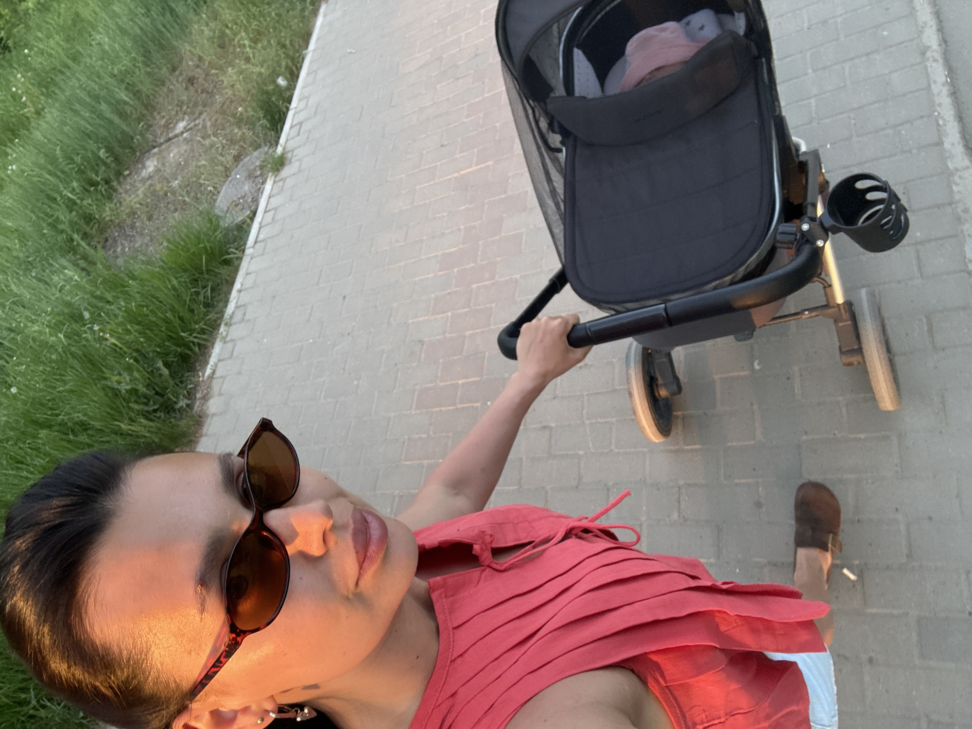 A person wearing sunglasses and a pink top pushes a baby stroller on a brick-paved walkway. The baby in the stroller is wearing a pink hat. The walkway is bordered by grass. A single brown sandal is visible on the walkway. The location is potentially in Chaikovsky, Russia.