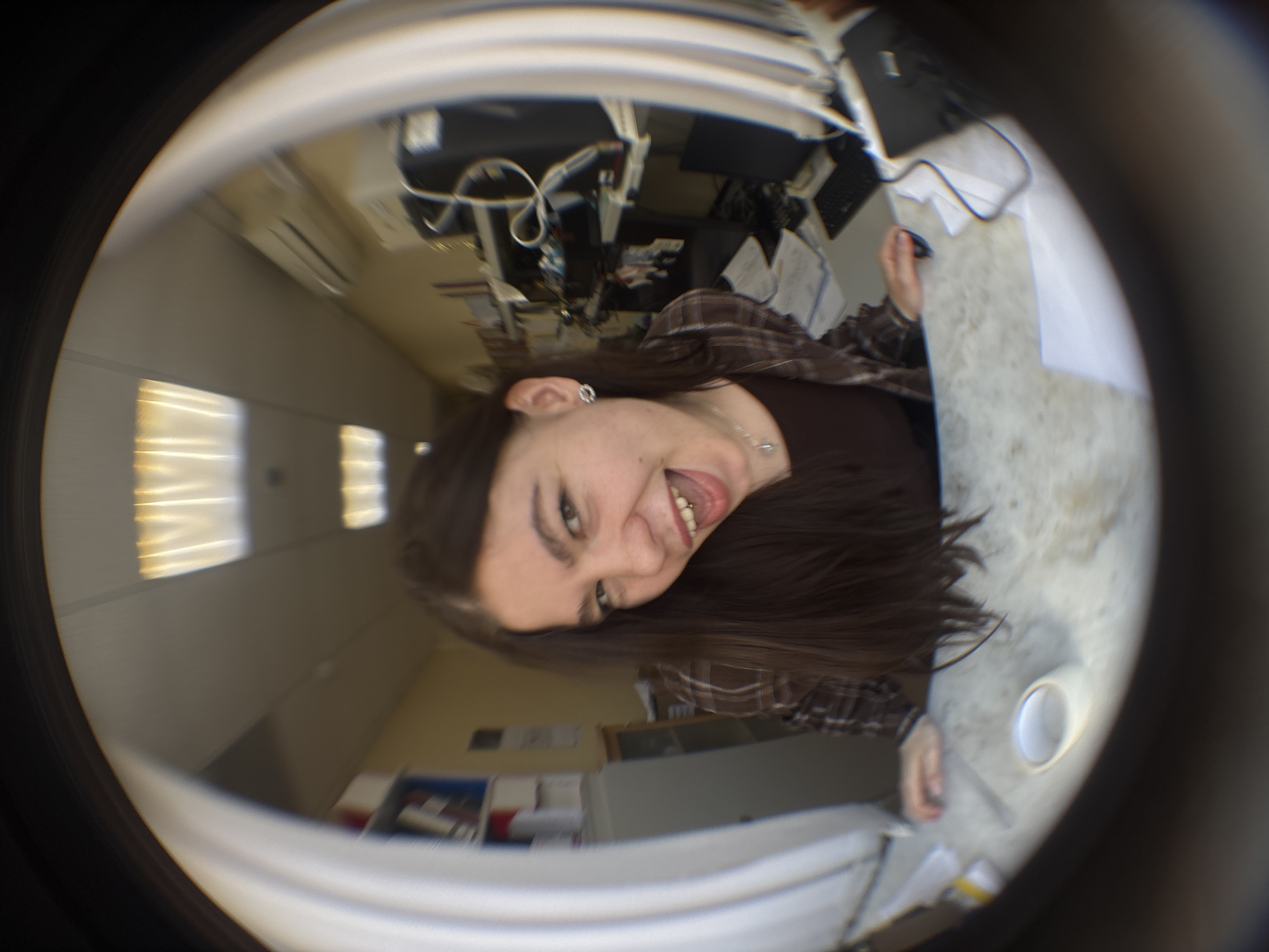 The image, taken with a fisheye lens, shows a woman sticking her tongue out at the camera. She has long brown hair and is wearing a brown plaid shirt. A white cup is on a gray surface. In the background, there is office equipment, including computers and cables. The location appears to be an office space, suggested by the fluorescent lighting and file cabinets. There is not enough information to determine the city of Perm, Russia.