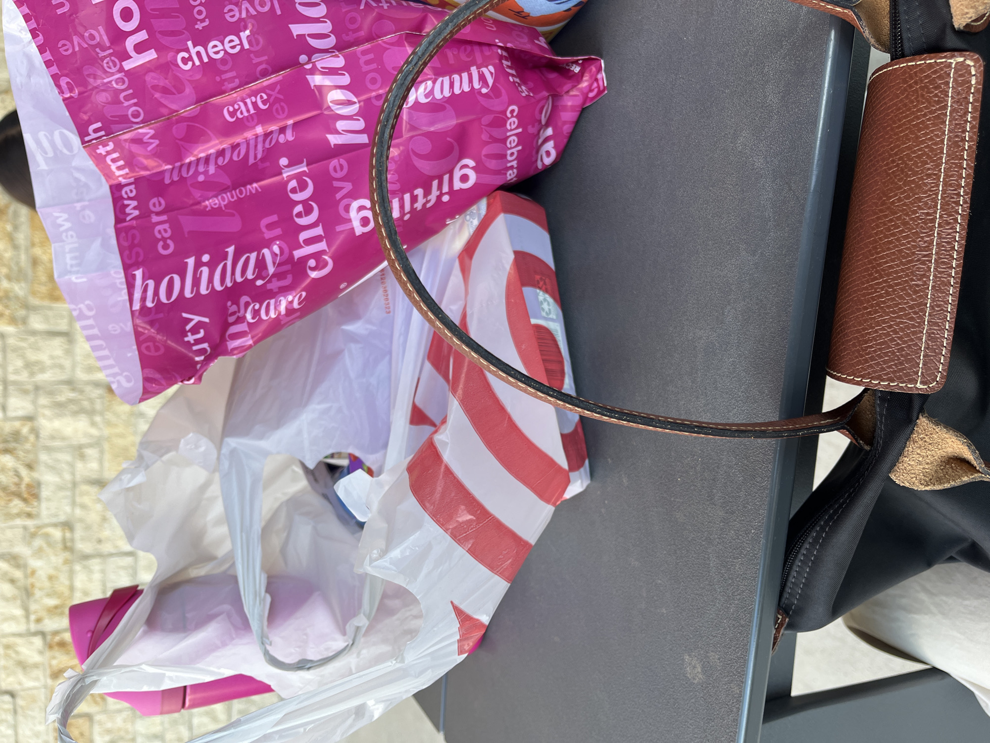 The image shows a collection of shopping bags and a black Longchamp tote bag resting on a gray surface, possibly a table or ledge. The shopping bags include a pink bag with the words "holiday cheer" printed on it, a Target bag with red and white stripes, and a white plastic bag, likely containing various purchases. The Longchamp tote bag is black with brown leather accents on the handle and flap. The photograph appears to be taken outdoors, with the corner of a paved surface visible in the background. The image shows items bought at North Star Mall in San Antonio.