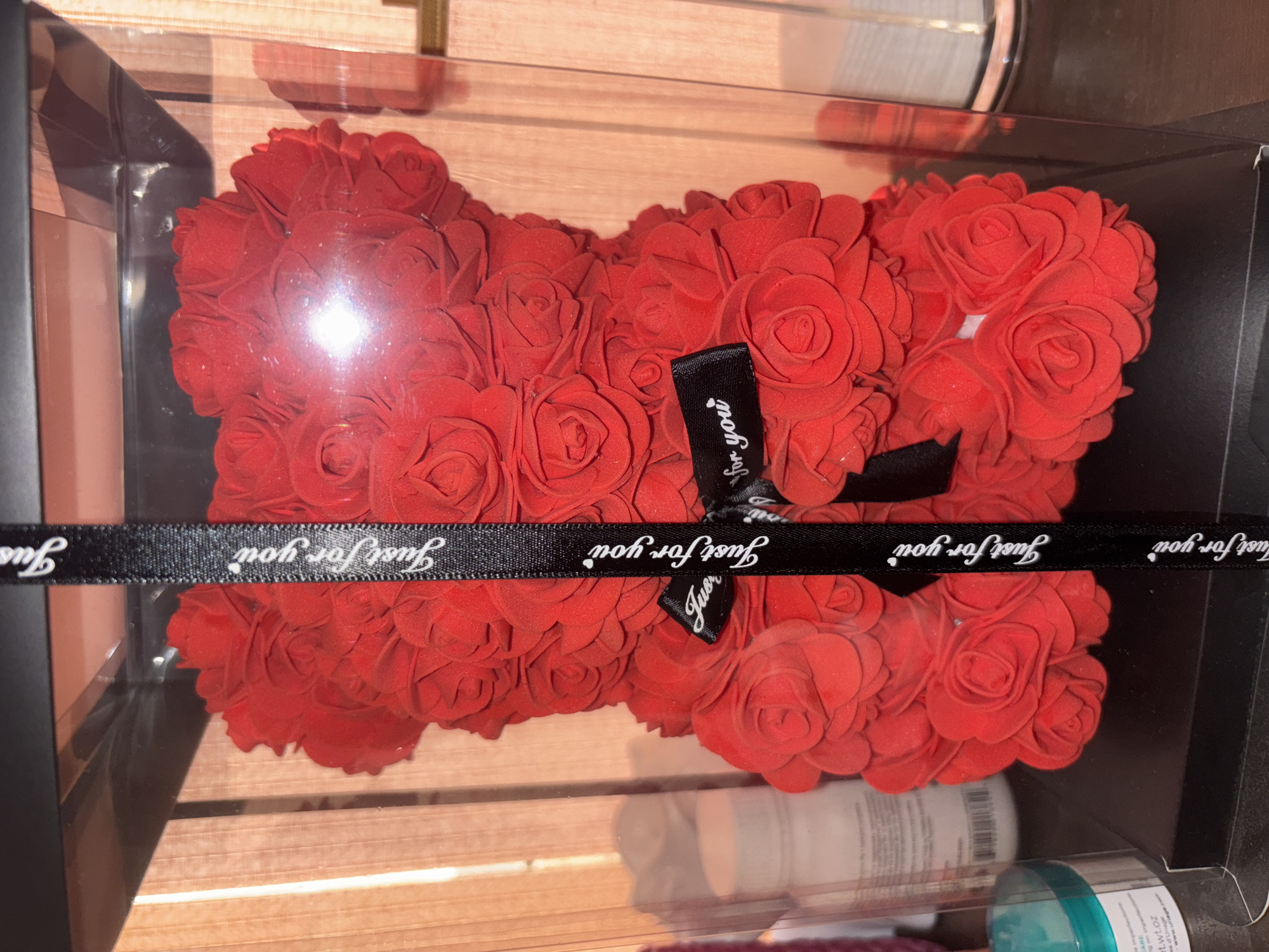 This media file is a close-up, top-down shot of a red rose teddy bear inside a clear box. The bear is made of many small red fabric roses. A black ribbon with white text wraps around the bear. The visible text on the ribbon reads "Just for you" repeated multiple times. The bear is positioned on a surface that appears to be made of light-colored wood or a similar material with horizontal lines. The lighting suggests an indoor setting, possibly a gift shop or a home. There are some reflections on the surface of the clear box, including a bright light source and what looks like the bottom of other objects or a shelf. Some small, partially visible cosmetic jars or bottles are also in the lower part of the frame, outside the box. The overall presentation indicates this is likely a gift item.