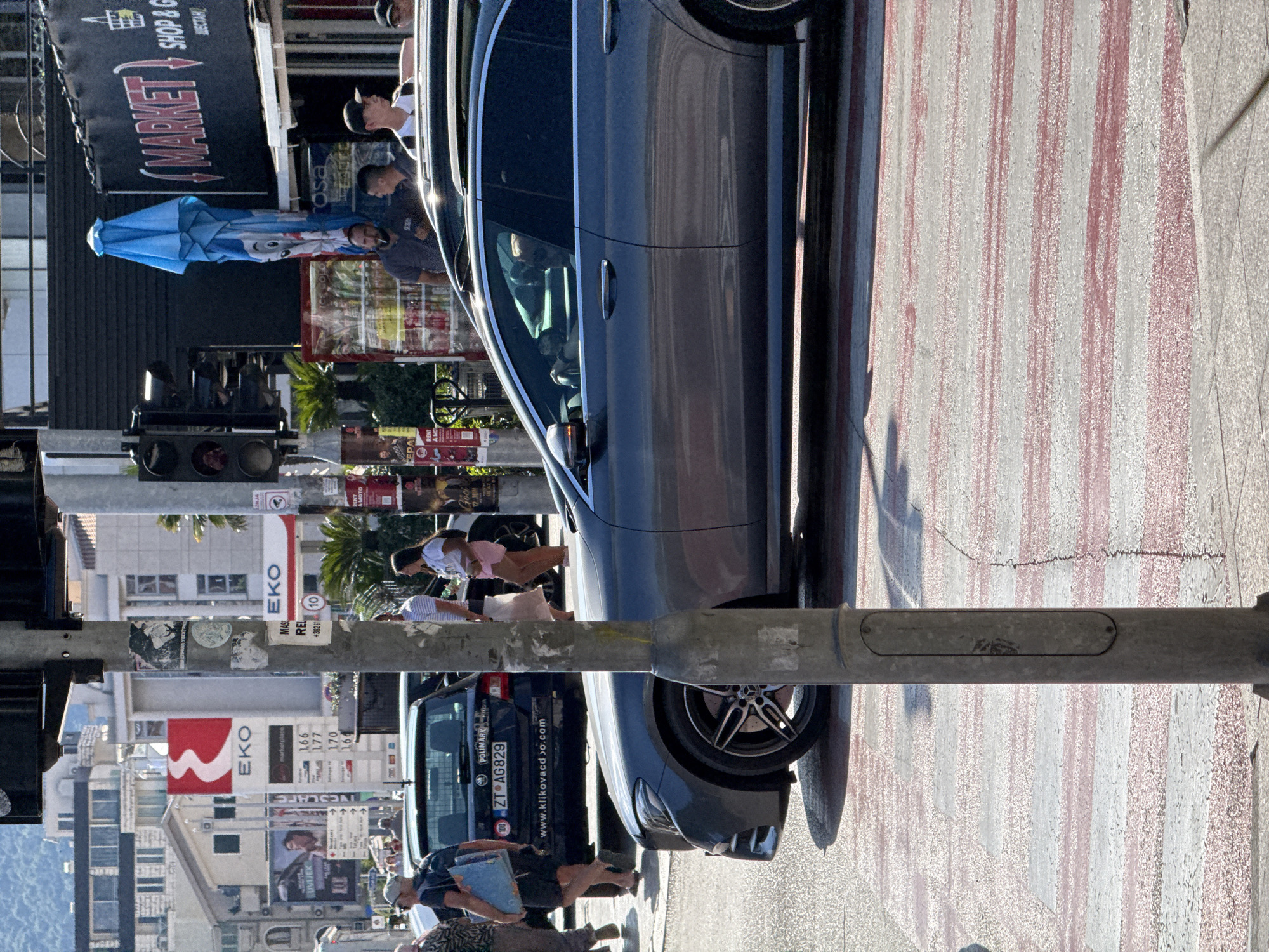 In Budva, Montenegro, on a sunny day, a busy street scene unfolds. A gray Mercedes sedan is stopped at a crosswalk, its driver visible behind the wheel. To the left, a dark blue Skoda is also parked, its rear license plate reading "ZT AG829". Pedestrians are walking on the sidewalk. In the background, buildings line the street, including a structure with an "EKO" gas station sign and another displaying "MARKET SHOP & G". Traffic lights and street poles are also present. The crosswalk itself is marked with red and white stripes.