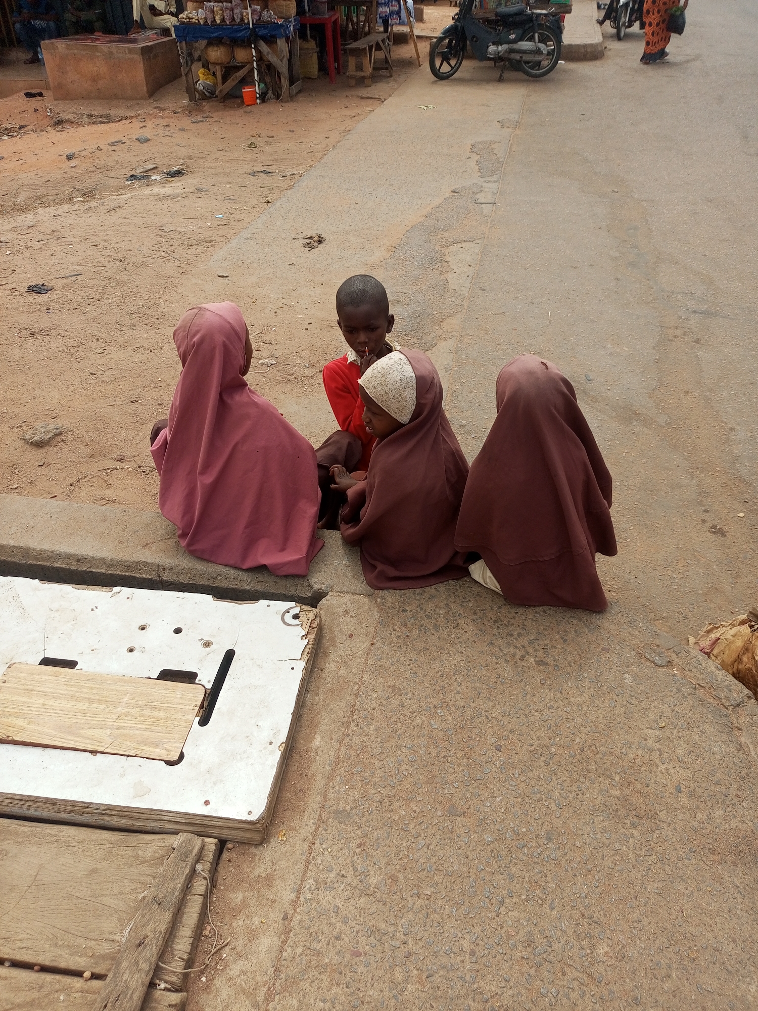 Three young children are seated together on a concrete curb. Two are wearing head coverings, while the one in the middle is not. The children are positioned near what appears to be a white, rectangular structure, possibly a tabletop or platform. In the background, a dirt road is visible, with a motorbike parked further down and a few small vendor stalls are set up in the distance. The setting is Tempure, Nigeria.