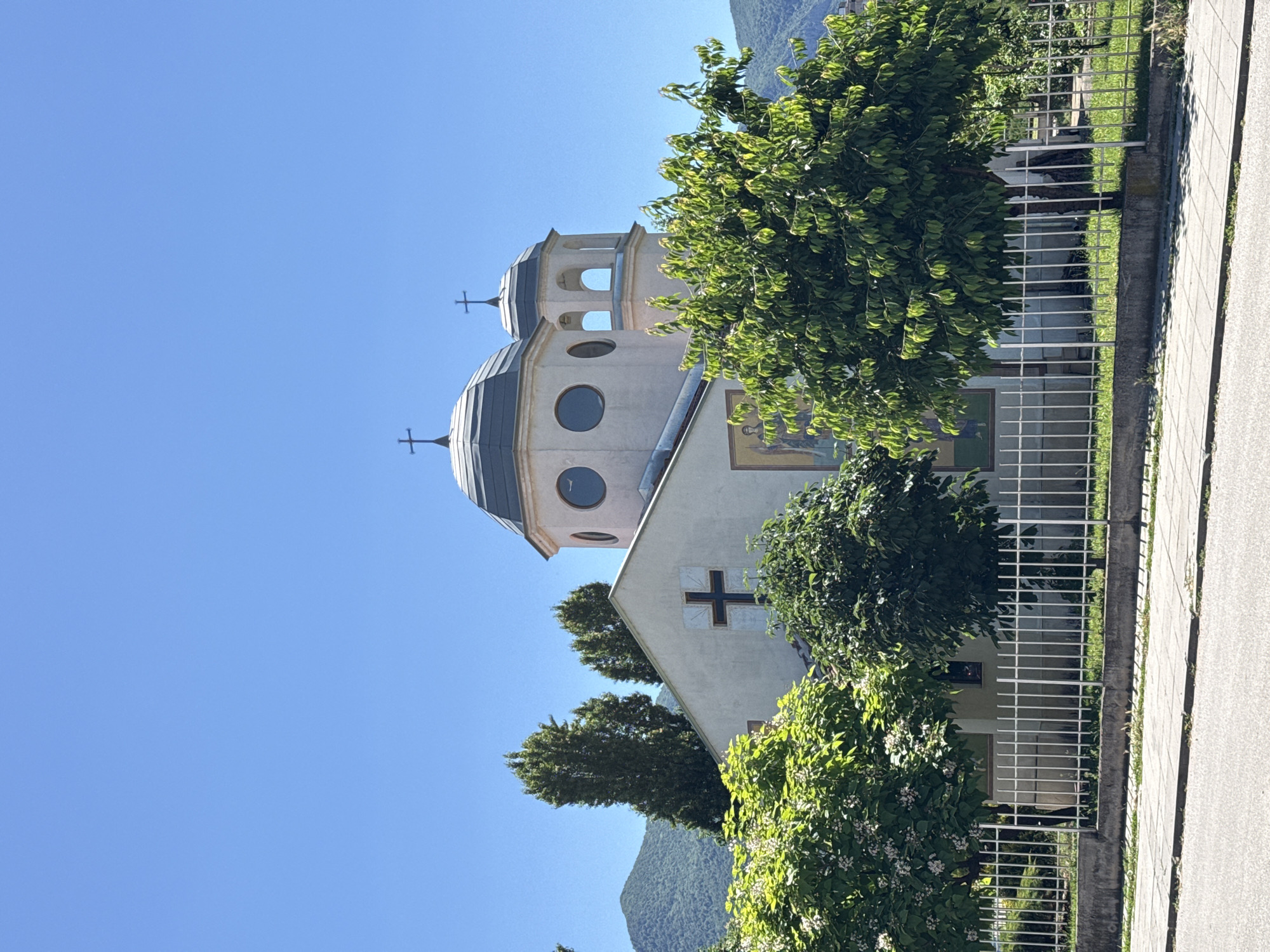 The image shows a church with two domes against a clear blue sky. The church is light-colored with a dark-domed roof. A cross is visible on the front of the building above a mural. Trees and foliage surround the church, and a white metal fence runs along the front. Mountains are visible in the background. The scene appears to be a serene day in a town or city. The location is in Vratsa, Bulgaria.