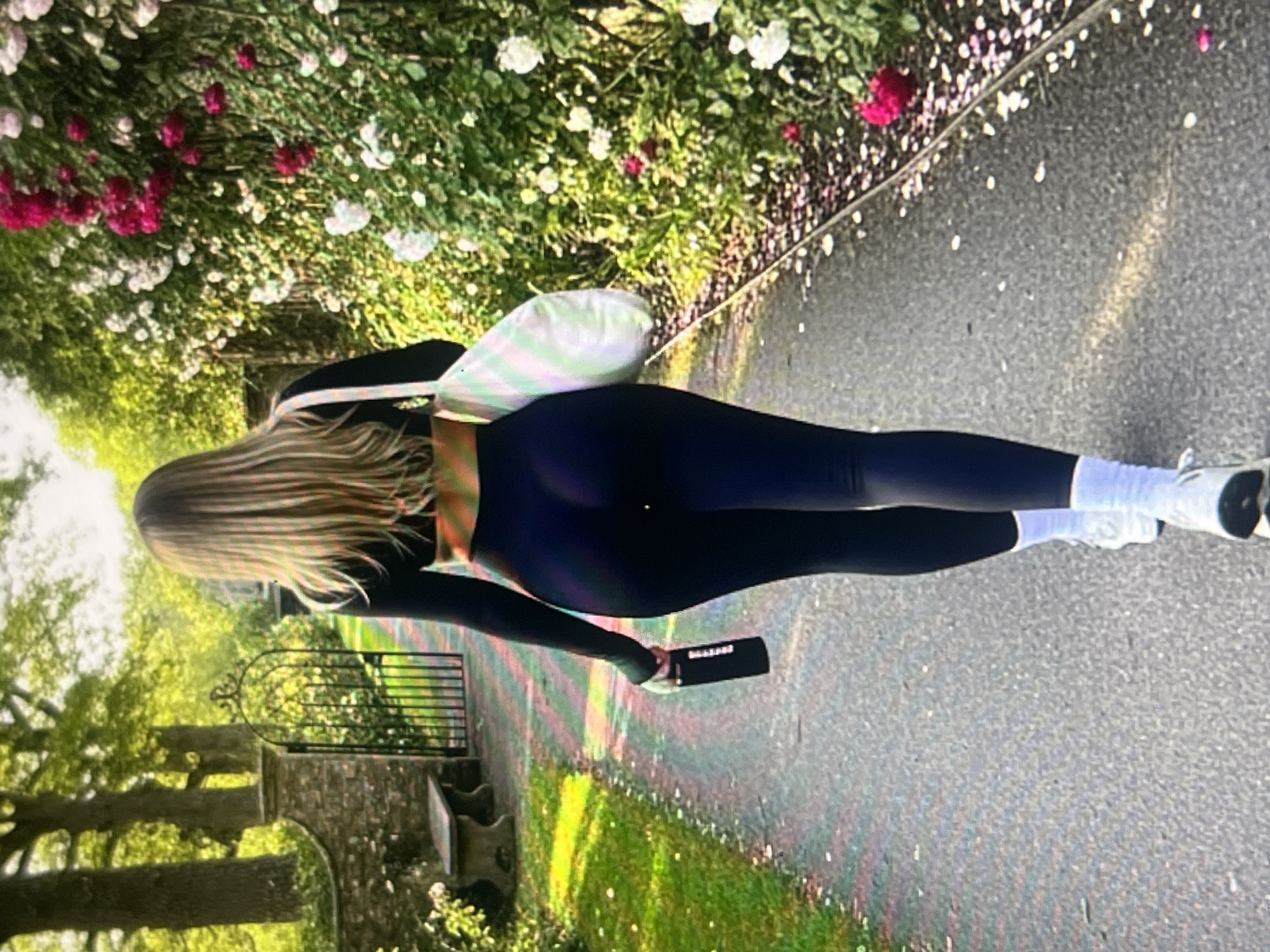 A woman walks away from the camera on a paved path lined with trees and flowers. She has long, blonde hair and is wearing a black crop top, black leggings, and white socks with white sneakers. She is carrying a white tote bag over her right shoulder and a black water bottle in her left hand. The path is surrounded by lush green grass and a stone wall on the left, with a black wrought-iron gate. On the right, a dense hedge of pink and white roses blooms, with some petals scattered on the path. The lighting suggests it is daytime, possibly late afternoon, given the warm glow on the grass.