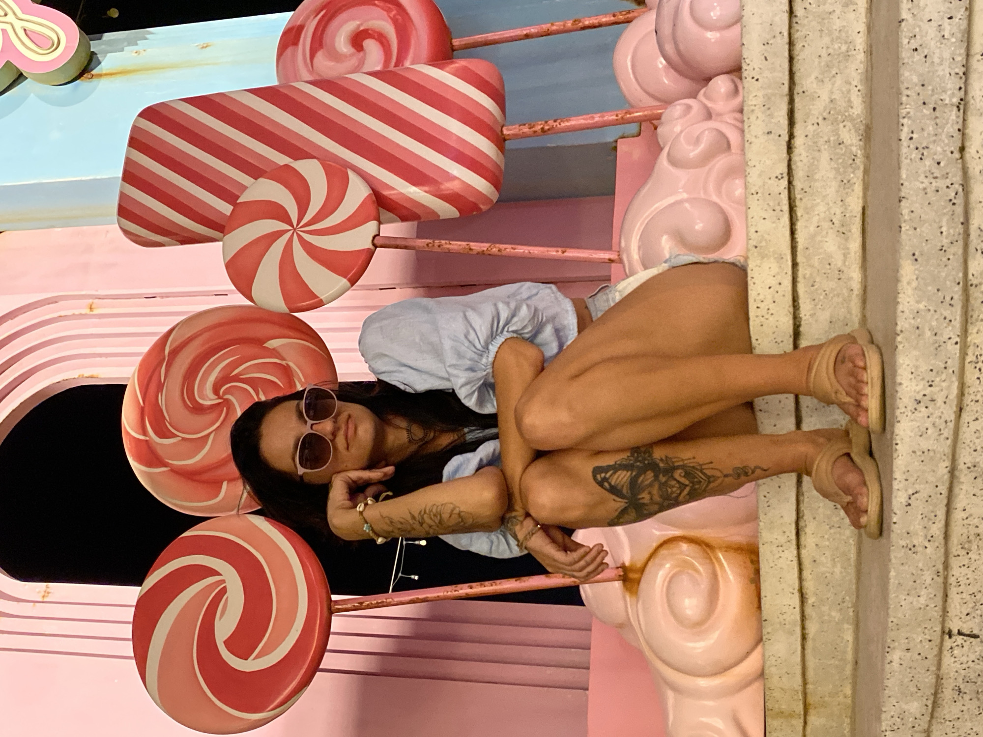 A woman with long dark hair and sunglasses sits on a tiered candy-themed structure. She is wearing a light blue puff-sleeved shirt and white shorts. Her left arm is resting on her knee, and her right arm is bent at the elbow. She has a butterfly tattoo on her right thigh. The background features oversized, colorful lollipops and candy shapes. The location is Tien Thanh Ward, Vietnam.