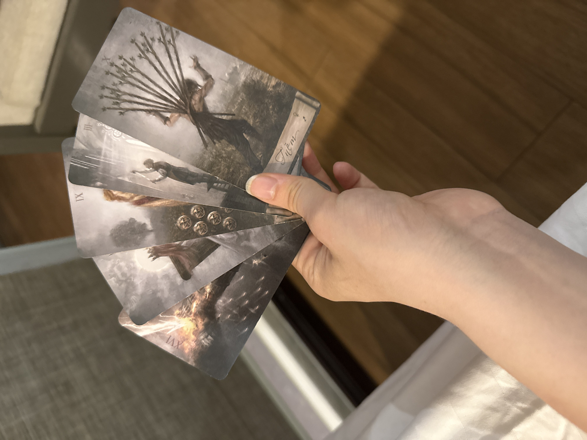 A hand holds a fanned stack of tarot cards. The cards depict dark, symbolic imagery. The top card shows a figure struggling against a bundle of sticks. Other cards depict figures in motion, coins, a phoenix-like bird, and a burning tower. The background is blurred and shows elements of a domestic interior. The image does not provide enough information to determine the location in Bac Nha Trang Ward, Vietnam.