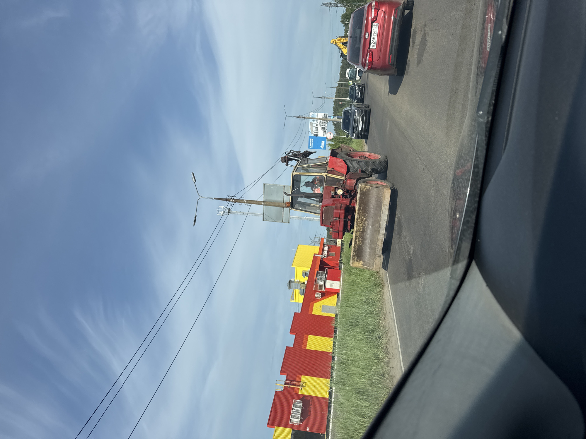 A daytime view from inside a car captures a red tractor with a front-end loader moving along a road, presumably in Chaikovsky, Russia, given the vehicle registration visible in the photograph. The tractor appears to be slightly ahead of a line of cars that include a red Kia. A man is visible in the tractor's cabin. A building with red and yellow sections stands beside the road. Overhead electrical lines stretch across the sky.