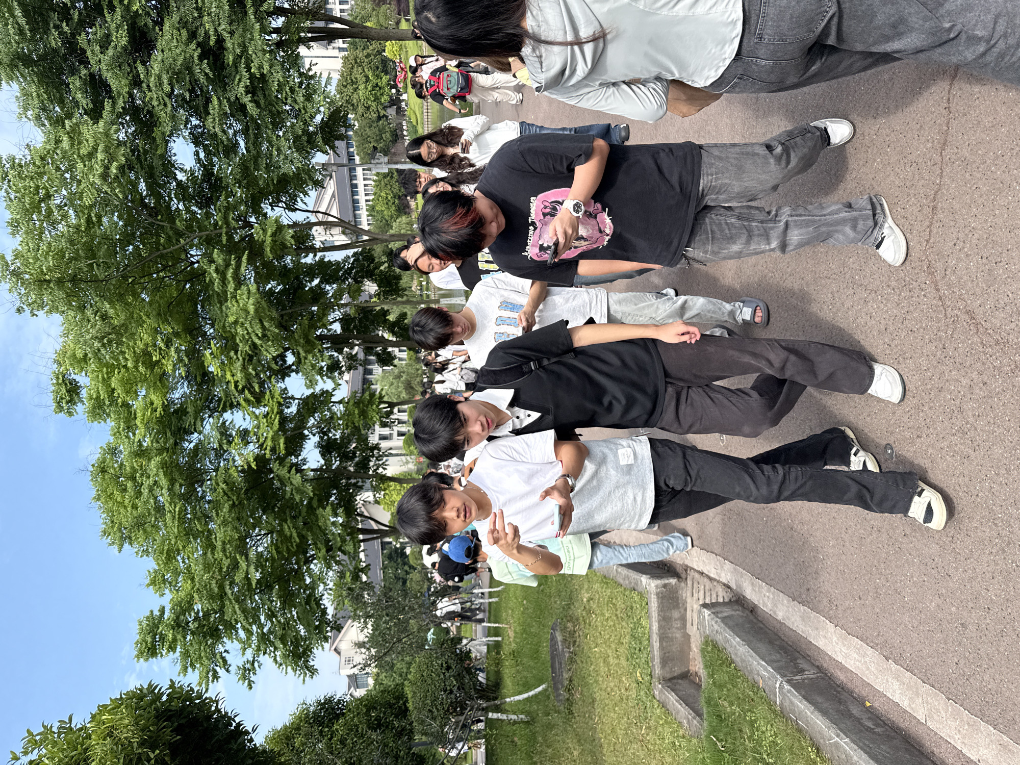 A group of young people are standing in a line on a paved pathway outdoors. The pathway is bordered by a low concrete wall and grassy area. Trees provide shade overhead. In the background, buildings are visible. The scene appears to be in a park or on a campus in Jiangjin, China.