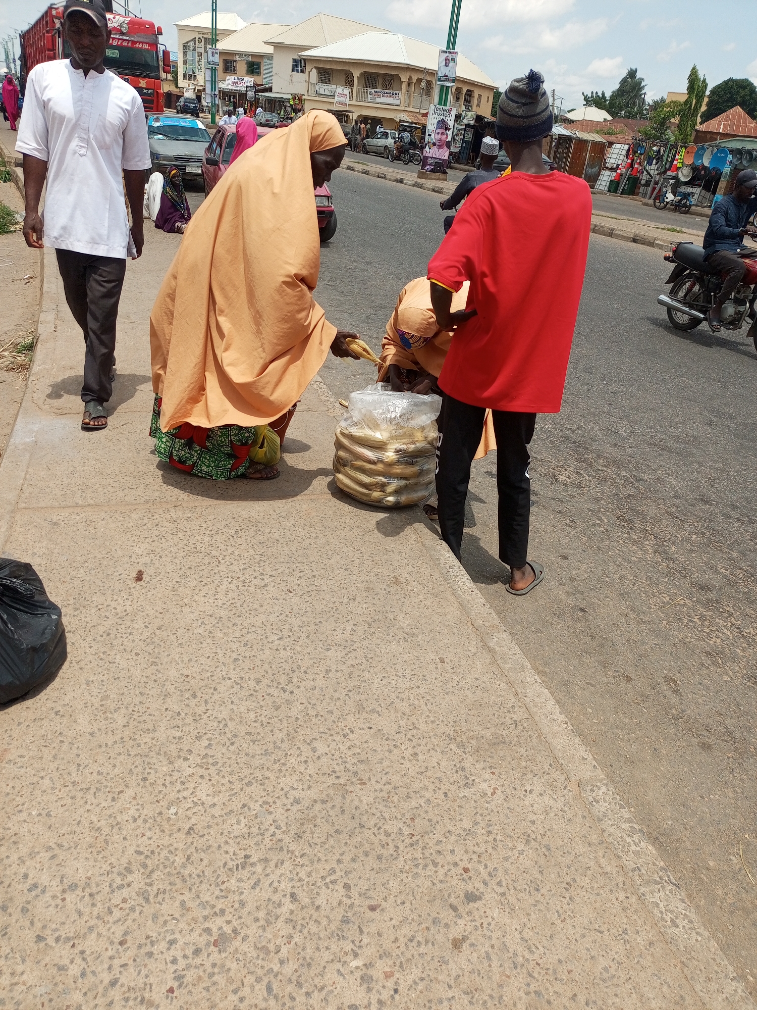 The image depicts a street scene in Arawa, Nigeria. In the foreground, a man wearing a white shirt and dark pants walks on a concrete sidewalk. To his right, a woman in a light orange hijab and a patterned skirt is interacting with a young man wearing a red shirt and dark pants, who is facing away from the camera. They appear to be exchanging items, with the woman holding what looks like corn. A large clear plastic bag filled with corn is on the ground between them. In the background, several buildings line the street, with some appearing to be shops. Vehicles, including a red truck, cars, and motorcycles, are visible on the asphalt road. The sky is overcast with some blue patches.