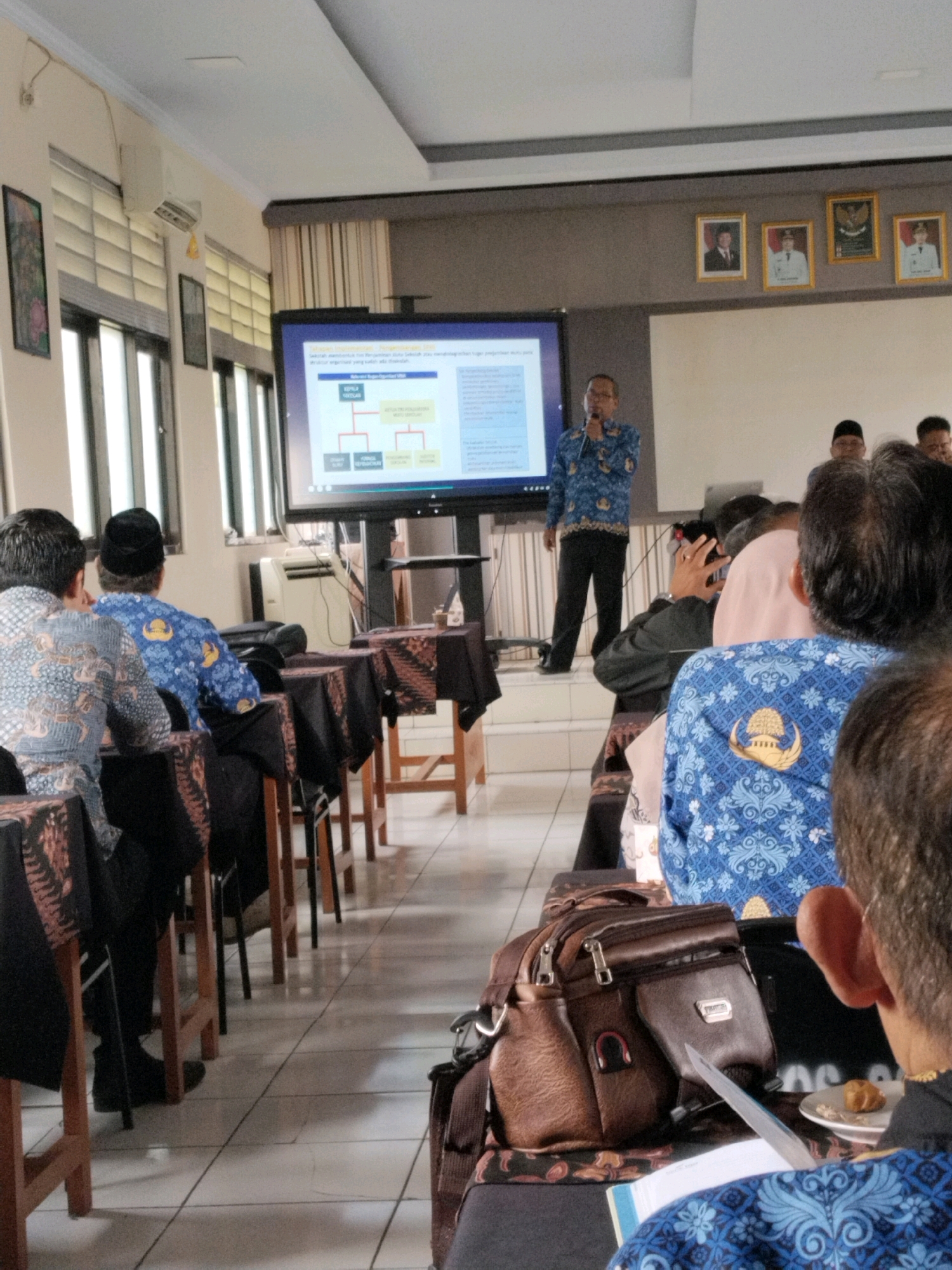 In a well-lit conference room, a presenter in a blue batik shirt and black pants stands on a small stage, holding a microphone and speaking into it. He gestures with his left hand as he addresses an audience seated at tables covered with patterned cloths. The presenter is positioned in front of a large screen displaying a flowchart. The room appears to be in a public building, possibly a government office or educational institution. The presence of national flags and portraits on the wall suggests an Indonesian context. The audience members are predominantly male and appear to be engaged in the presentation. One person in the foreground is looking down at a document, while others are looking towards the presenter. The overall atmosphere is one of a formal presentation or training session.