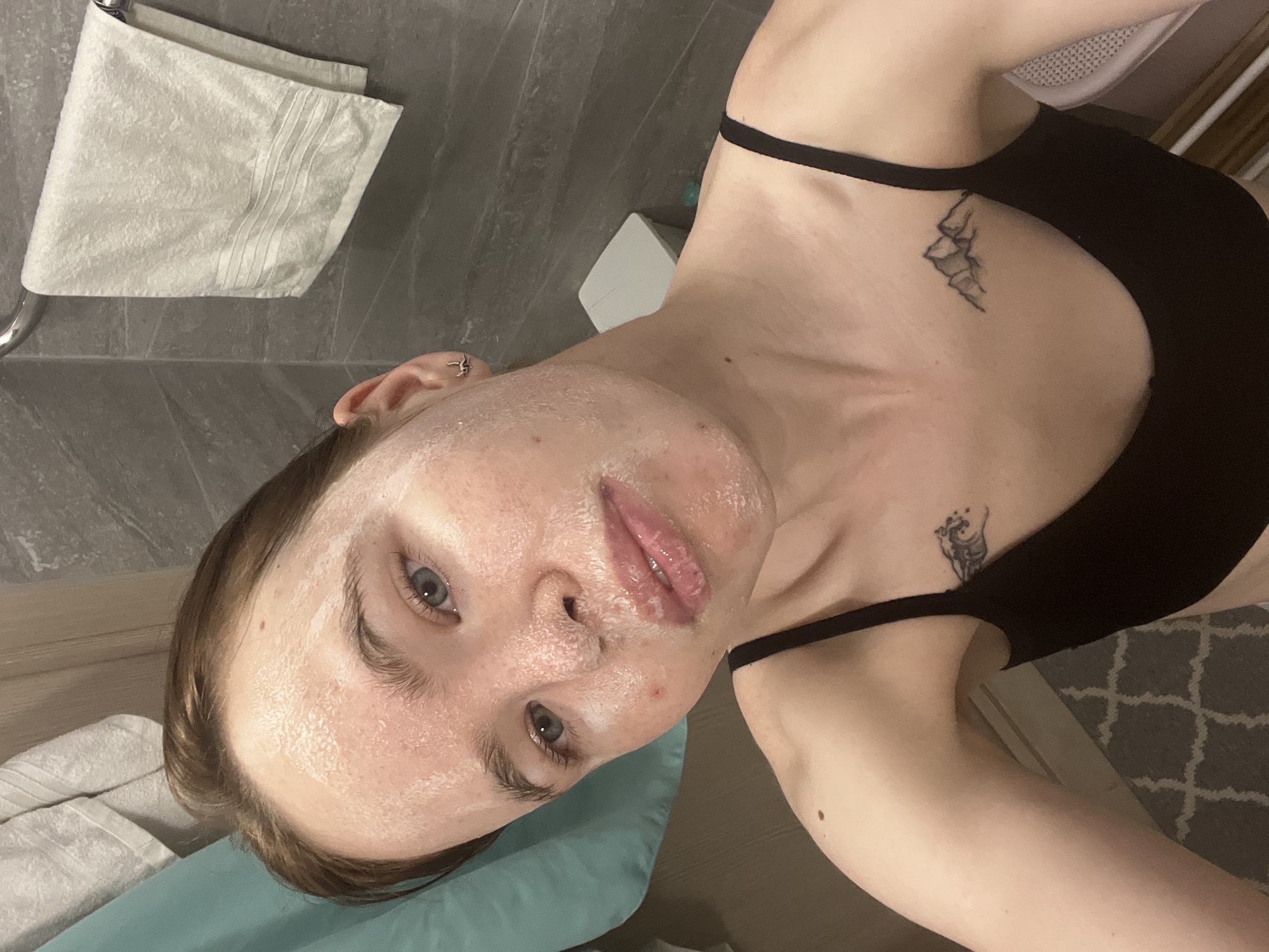 A close-up, selfie-style photograph shows a woman with fair skin, partially covered in a white substance, possibly a face mask, from the forehead to her lips. She has light-colored eyes and visible pores. Her hair appears to be pulled back. She is wearing a black tank top, and there are two tattoos visible on her upper chest area.
The background suggests a bathroom setting, with a gray tile wall partially visible behind a white towel hanging on a metal bar. A light blue or green pillow is under her head. The bottom right corner reveals a light-colored patterned carpet or rug. The overall lighting is soft.
The city location is specified as Perm, Russia.
