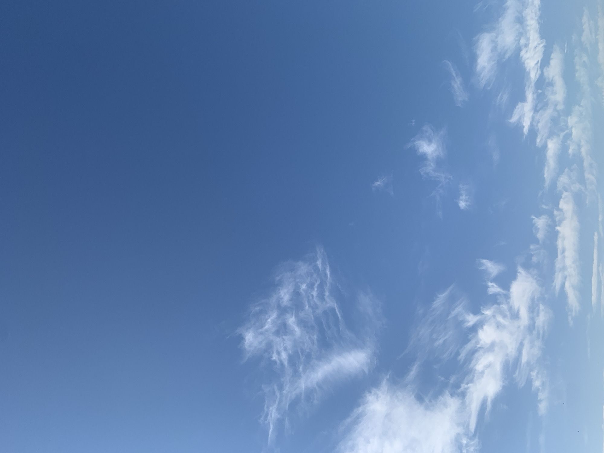 A high-angle, full-frame shot captures a bright blue sky with wispy, white clouds. The clouds are scattered across the frame, with a larger, more defined formation in the upper left quadrant and smaller clusters towards the bottom. The image appears to be taken on a clear day, possibly in Kolomyia, Ukraine, although no specific landmarks are visible. No people, objects, or actions are depicted in the scene.