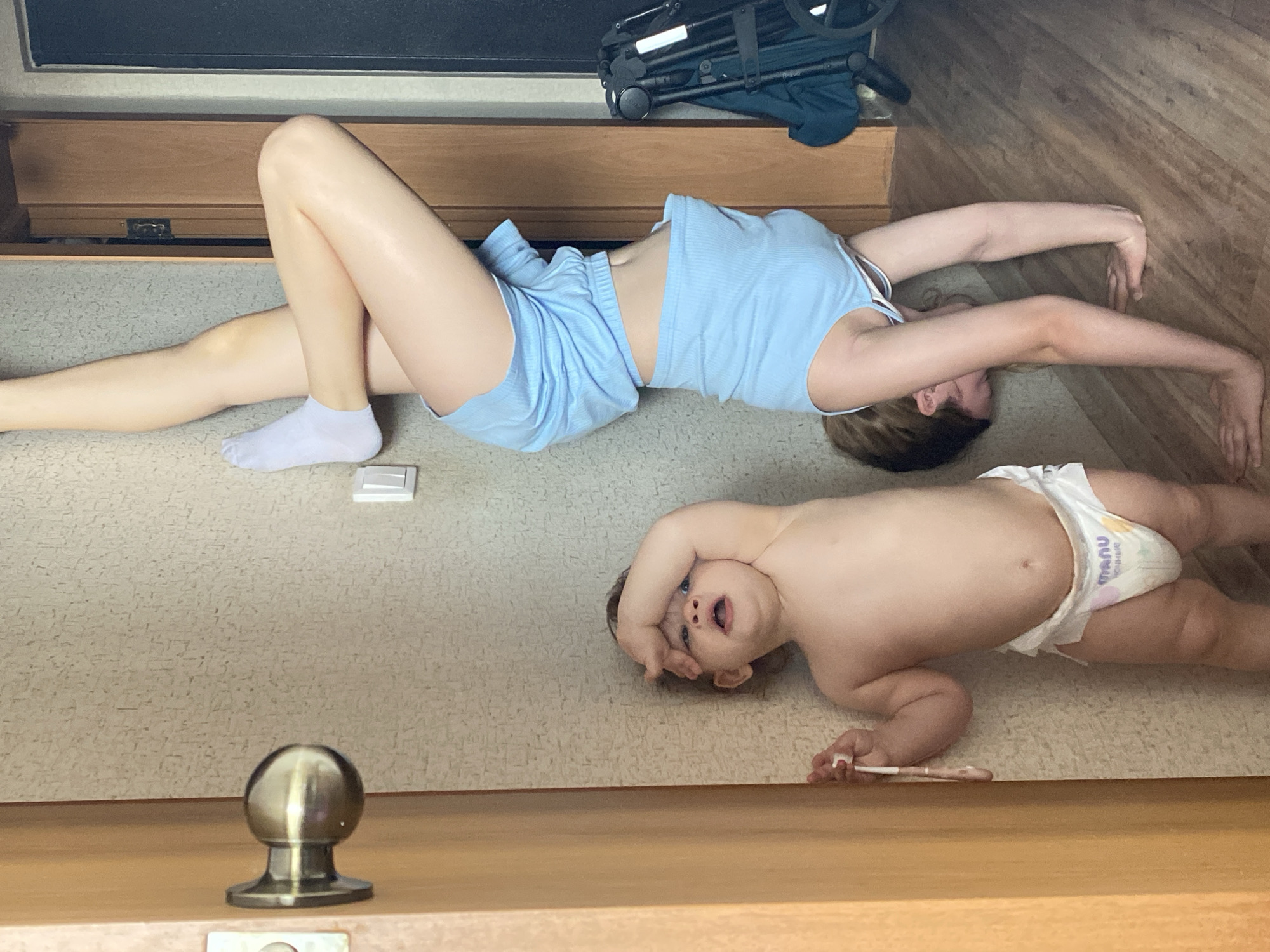 A high-angle shot shows a woman and a baby lying on their backs on a beige carpet in an indoor setting. The woman, wearing a light blue pajama set and white sock on her left foot, is contorted with her back arched and arms stretched out, reaching toward the right side of the image. The baby, wearing a diaper, is positioned below the woman. The baby's left hand is covering its face. A white object is positioned near the woman's left leg. In the background, there is a dark surface behind a wooden cabinet or drawer. A stroller is positioned on the cabinet, behind the woman. A round metal doorknob is in the bottom left corner. The location is Unknown, Unknown.