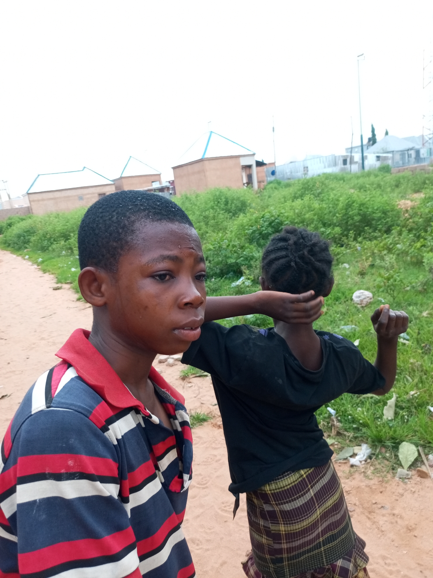 In Tempure, Nigeria, two young individuals, one in the foreground and another behind, stand on a sandy path. The individual in the foreground, facing left and appearing slightly distressed, wears a striped polo shirt with red, blue, and white bands and a red collar. Their face is moist, suggesting sweat or tears, and they have short, dark hair. The second individual, seen from the back, wears a black t-shirt and a plaid skirt. This person's left arm is raised, holding the back of their neck, while their right hand is clenched into a fist. Behind them, a patch of dense green vegetation lines the path, and in the distance, several buildings with light-colored walls and bluish roofs are visible, along with some light poles and what appears to be a tower. The sky is overcast.