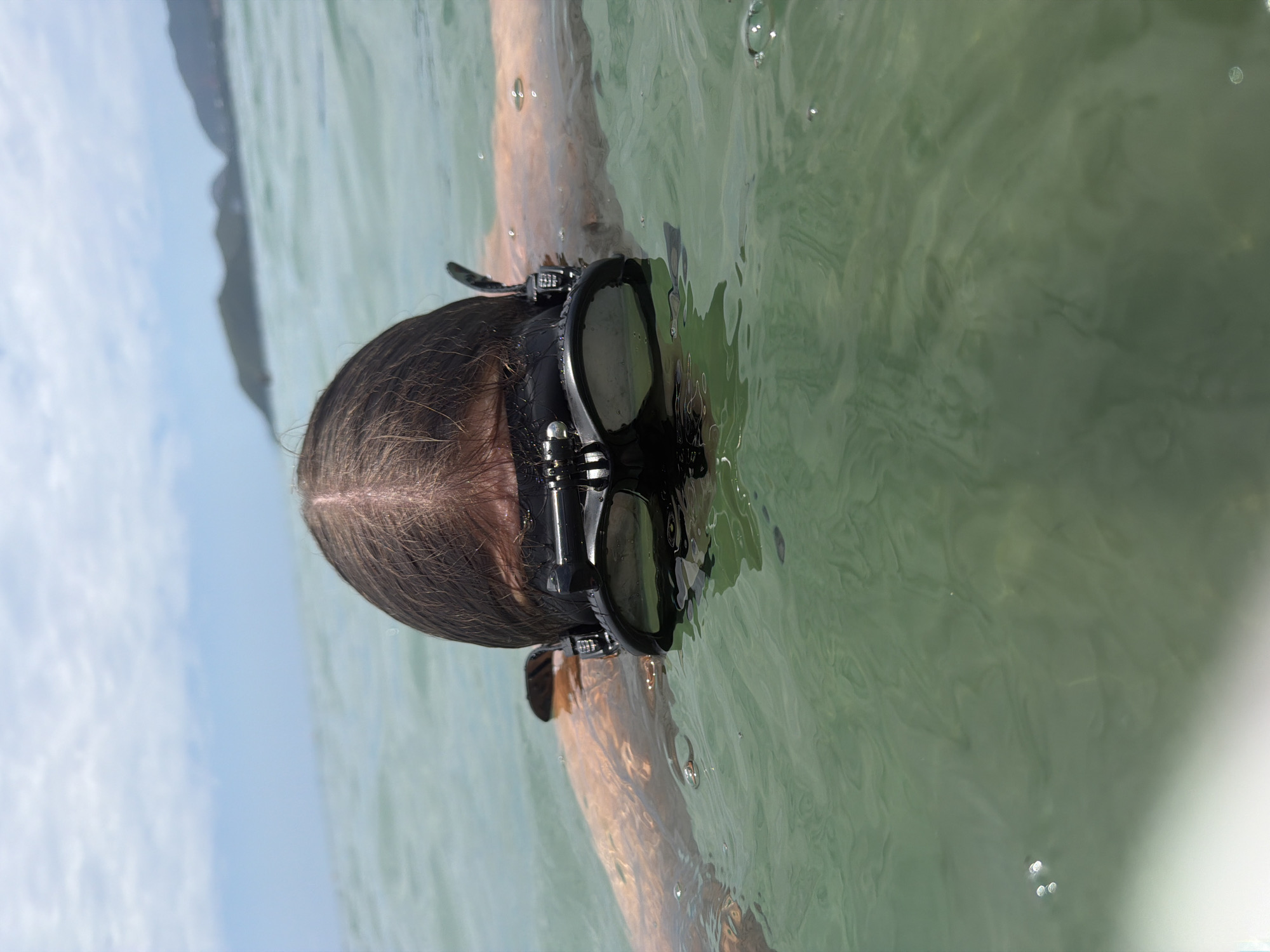 A person wearing a black mask with a GoPro attached is submerged in the ocean near a shore. The water is clear and green, with gentle waves. In the background, mountains are visible on the horizon under a hazy sky. The scene is likely in Nha Trang, Vietnam, a coastal city known for its beaches and water activities. The person appears to be swimming or snorkeling, with their head and shoulders above the water, and their body submerged. The GoPro is positioned on the mask, suggesting the person is recording their underwater experience. The lighting suggests it is daytime.