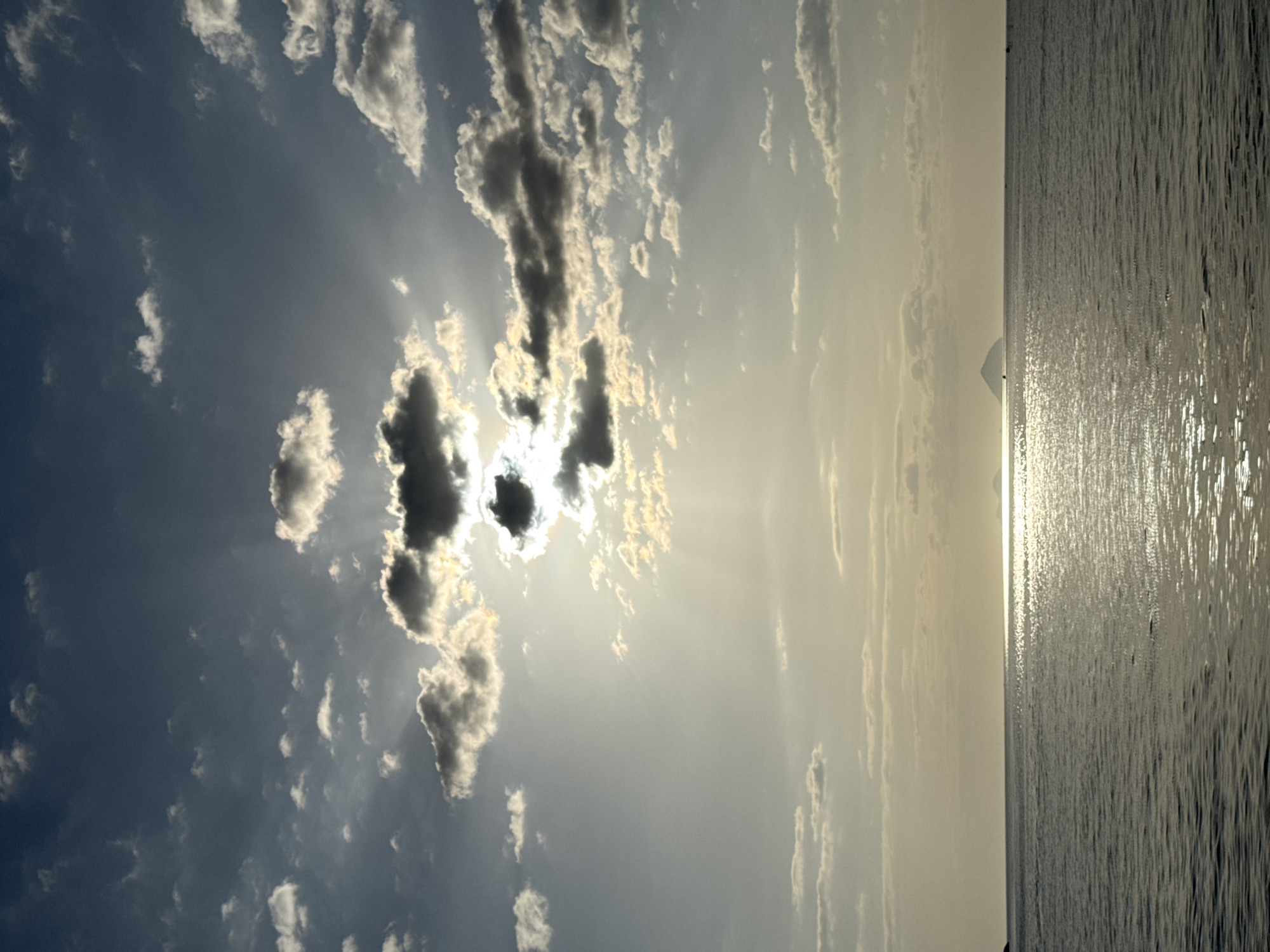 A wide-angle, horizontal, low-angle, eye-level shot captures a serene seascape with a dramatic sky. The sun, partially obscured by dark, wispy clouds, casts a bright, shimmering reflection across the calm ocean surface. The sky is a gradient of deep blue at the top, transitioning to a hazy, pale yellow near the horizon, with scattered white and grey clouds adding texture. In the distance, a small, dark island with a conical shape is visible on the right horizon, faintly illuminated by the setting sun. The water is a dark grey with subtle ripples reflecting the light. The scene appears to be taken from a beach or a boat in Nha Trang, Vietnam, at dusk. No people or specific structures are identifiable.
