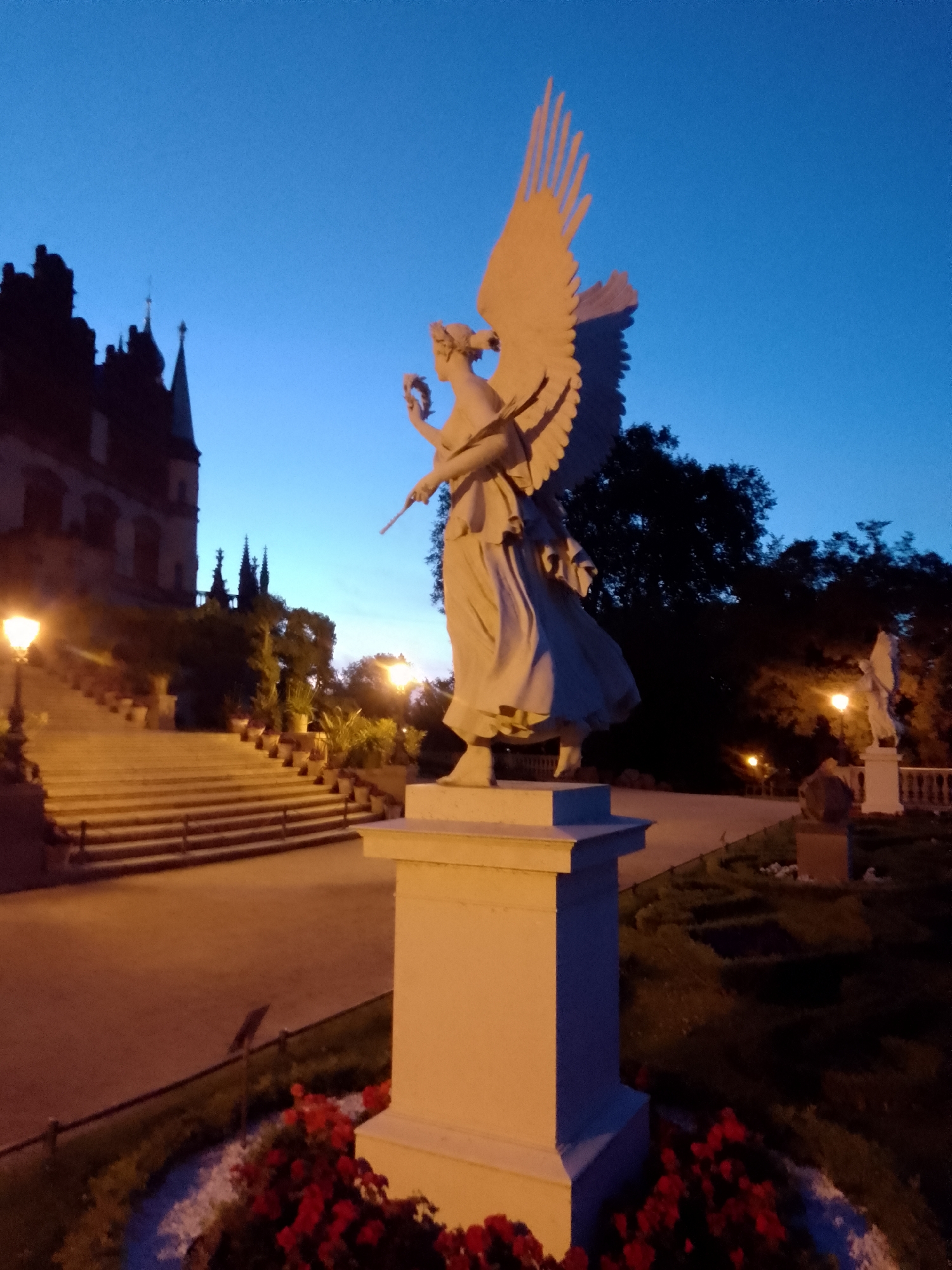 This media file captures a twilight scene at dusk, possibly in Schwerin, Germany, featuring an angel sculpture prominently in the foreground. The sculpture, made of a light-colored material, depicts an angel with large wings spread upwards, holding a laurel wreath and a staff. It stands on a pedestal in a garden setting.
Behind the angel sculpture, a grand staircase lined with potted plants leads up to a dark, imposing building, likely a castle or palace, with Gothic architectural elements. The sky is a deep blue, transitioning from lighter shades near the horizon, indicating the end of daylight. Several lampposts are illuminated, casting a warm glow on the staircase and pathway, creating an atmosphere of calm and serenity.
In the background, silhouetted trees frame the scene, and another, smaller statue can be seen in the distance, also illuminated by a lamppost. The grounds appear to be a well-maintained garden with flower beds, some of which are visible with red flowers in the foreground.
The overall mood is peaceful and somewhat majestic, with the sculptures and architecture suggesting a place of historical significance. The lighting and the darkening sky contribute to a tranquil evening ambiance. No visible text is present in the image.