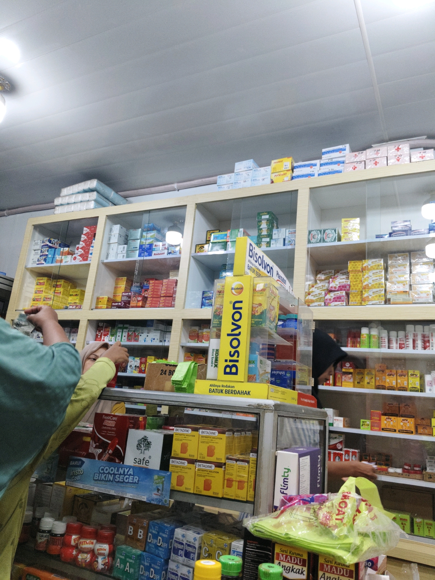 The image depicts the interior of a pharmacy in Lhokseumawe, Indonesia. The scene is dominated by shelves stocked with various medicines and health products. In the foreground, two individuals are visible. One, wearing a green shirt and headscarf, appears to be a customer making a purchase, holding what looks like cash. Another person, wearing a dark head covering, is partially visible behind the counter, seemingly assisting the customer.
The shelves are organized with boxes and bottles of medication, including brands such as "Bisolvon", "Insto", "Coolnya", and "Betadine". Several stacks of white packaged goods, possibly tissues or medical supplies, are placed on the top shelf. A prominent yellow display stand for "Bisolvon" is positioned on the counter. A bright green plastic bag filled with items is also on the counter. The lighting in the pharmacy appears to be artificial, coming from overhead fixtures. The overall environment suggests a typical retail setting for a pharmacy, with an emphasis on product display and customer service.