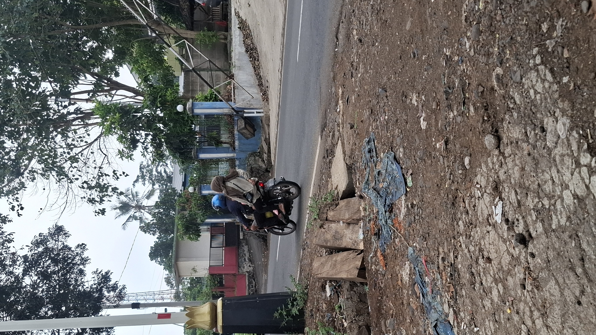 A medium shot shows two individuals riding a motorcycle on a street in Cilaku, Indonesia. The person in the front is wearing a blue helmet and a dark jacket, and the person in the rear is wearing a tan headscarf and a light-colored jacket. The motorcycle is moving away from the camera, with the rear passenger's back turned to the viewer. They are traveling on an asphalt road, with the left side of the road showing a dirt shoulder littered with debris, including pieces of wood, plastic sheeting, and rocks. The right side of the road features a retaining wall and a fence, with trees and buildings in the background. The sky is overcast, suggesting a cloudy day.