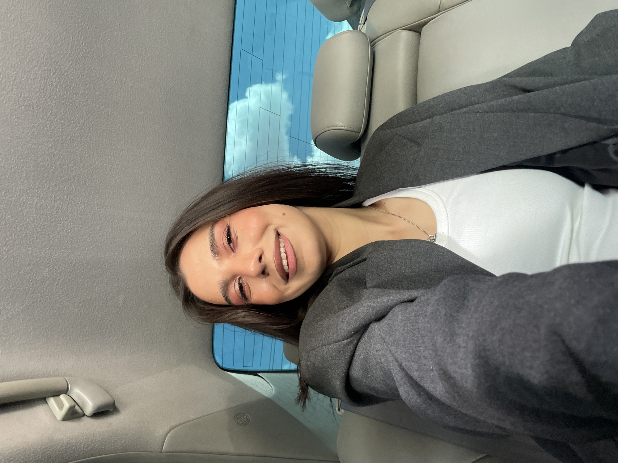 A young woman is seated inside a car. She is wearing a white top and a gray jacket. She is smiling. The interior of the car is gray, and through the car window, a view of the sky with clouds can be seen. The location is not possible to identify as Yerevan, Armenia with the given data.