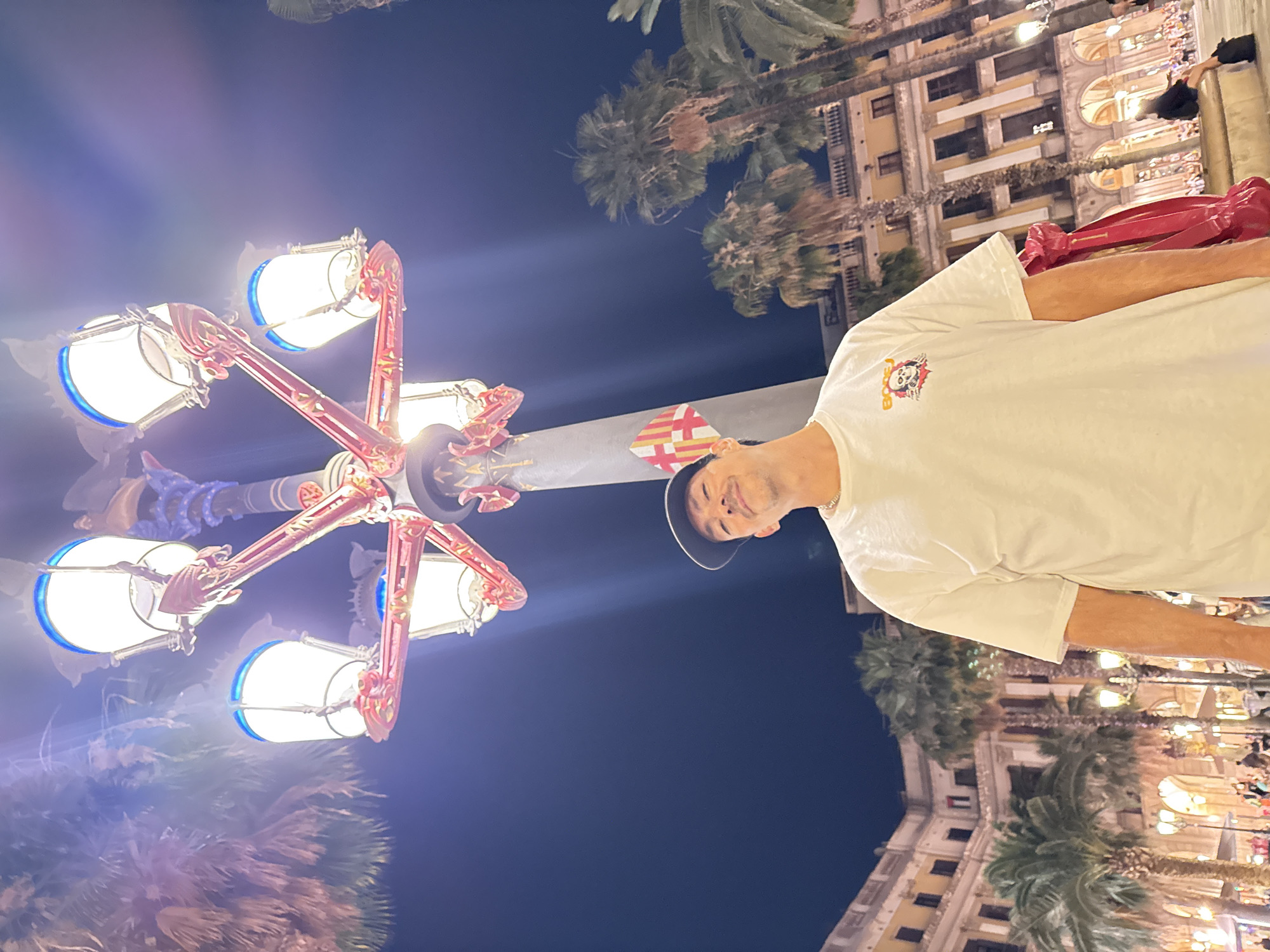 A medium shot shows a man wearing a white t-shirt and a baseball cap, posing under a decorative street lamp at night. The lamp has multiple lights with blue accents and red ornate detailing. Behind the man, palm trees and buildings with illuminated windows are visible, suggesting an urban setting with a warm climate. The overall atmosphere is lively, with blurred figures and lights in the background indicating a public square or street scene in Barcelona, Spain, possibly Plaça de Catalunya given the architecture and street lamps. The man's expression is a slight smile.