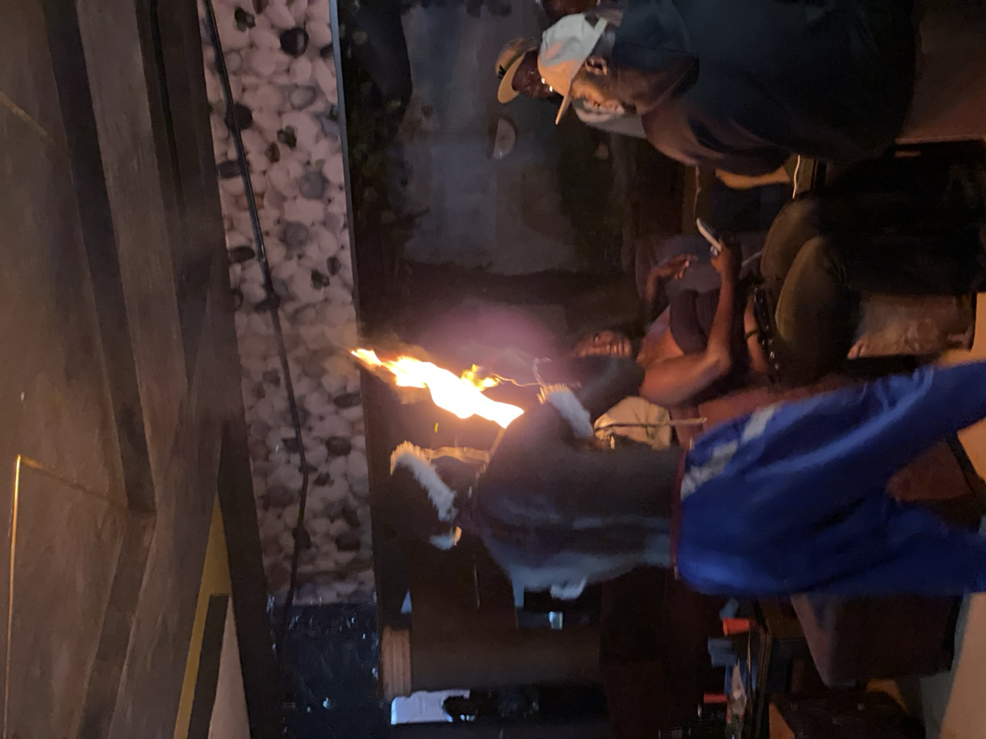 In Durumi, Nigeria, a performer with a white headdress and what appears to be horns is seen from the back, holding a flaming torch in their right hand. The performer wears a dark top and loose blue pants with gray accents. To the right of the performer, a woman in a black strapless top and dark pants is seated, looking towards the performer. Further to the right, two men are visible; one wearing a light-colored baseball cap and a dark hoodie, and another in a tan hat and glasses. The scene takes place indoors, with a textured stone-like wall and a ceiling with visible beams. Several red cups are on a table in the background, to the left of the performer. The lighting is dim, with the primary illumination coming from the torch.