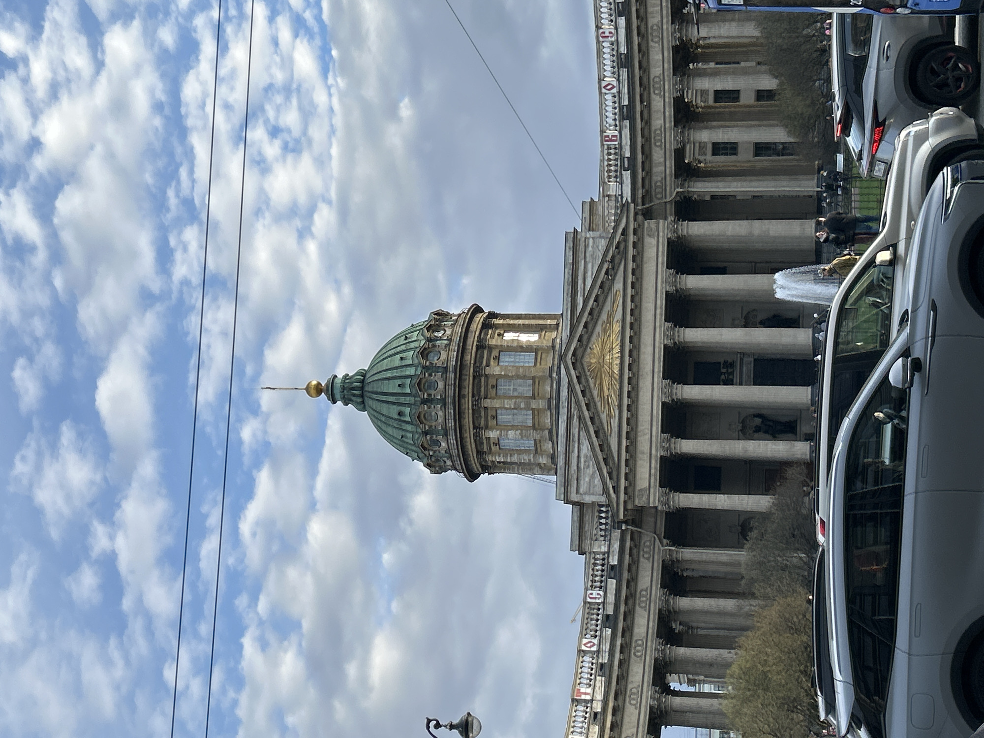 The vertically oriented image displays a partial view of the grand Kazan Cathedral in Saint Petersburg, Russia, under a bright, partly cloudy sky. The most prominent feature is the cathedral's distinctive green-patinated dome, crowned with a golden finial, resting on a multi-tiered drum with numerous windows. Below the dome, a classical facade is visible, featuring a triangular pediment adorned with sculptural relief, supported by a prominent colonnade of imposing columns. A balustrade above the columns features decorative red and white elements, including what appear to be small banners or crests with white symbols, possibly Cyrillic letters or crosses, on a red background.
In the foreground, a city street runs parallel to the cathedral, with several cars, mostly light-colored SUVs and sedans, either parked or moving, partially obscuring the lower sections of the building. A small fountain, actively spraying water, is visible between the cars and the cathedral's entrance. A few people can be seen on the sidewalk or near the fountain, including one person in a dark jacket. Overhead, two dark utility lines stretch across the blue sky, which is dotted with scattered white cumulus clouds. A classic black streetlight is partially visible in the lower left corner. The scene depicts a lively daytime urban environment centered around the iconic architectural landmark.