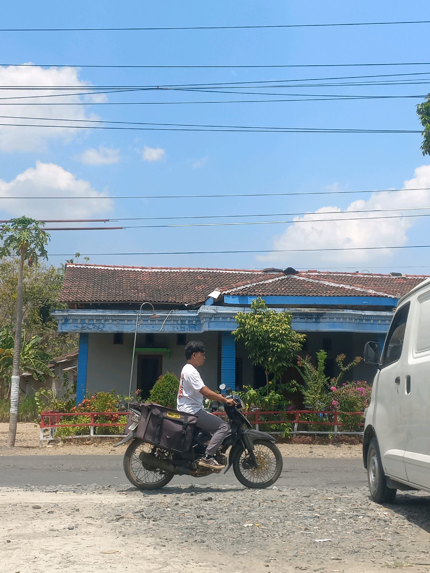 A young man wearing a white t-shirt and grey pants rides a black motorcycle with saddlebags on a paved road. He is seen from the side, looking forward as he rides. The motorcycle has a gold-colored rim on the front wheel and a grey saddlebag on the rear. To the right of the motorcycle, the side of a white van is visible. Behind the motorcycle, a house with a red tile roof and blue trim is situated. The house has several windows and is surrounded by green trees and flowering bushes. A tall, thin tree with white paint on its lower trunk stands to the left of the house. Overhead, power lines are strung across a bright blue sky with scattered white clouds. The ground in the foreground is covered in gravel and dirt. This scene appears to be in the rural outskirts of Baturetno, Indonesia, given the architectural style of the house and the presence of vegetation common to the region.