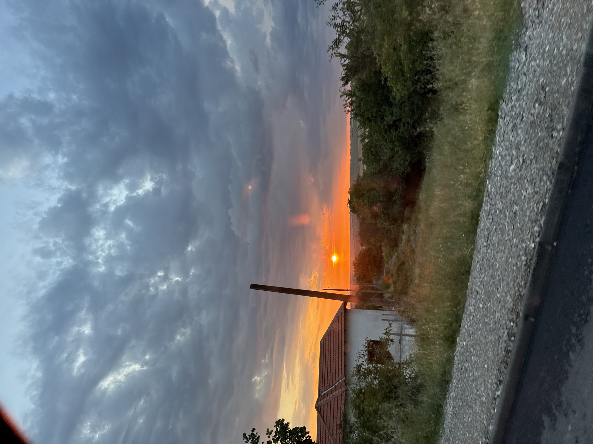 The image depicts a sunset over a rural landscape near the city of Probuda, Bulgaria. The sky is filled with dramatic, dark clouds, with patches of vibrant orange and yellow light breaking through near the horizon where the sun is setting. In the foreground, a gravel road borders a paved surface, likely a street or highway. To the left, a white house with a terracotta tiled roof is partially visible, surrounded by overgrown grass and bushes. A tall, slender wooden pole stands near the house, silhouetted against the colorful sky. The overall scene captures a serene yet somewhat dramatic natural event in a semi-rural setting. There are no people or animals visible in the image.
