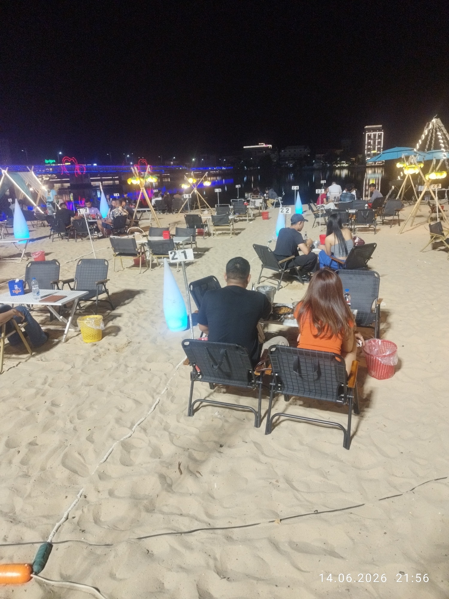 The image depicts an outdoor dining area on a sandy beach at night. The scene is set along the Kampot River in Kampot, Cambodia, with the river and buildings illuminated in the background. Several groups of people are seated at tables, enjoying their meals and drinks. Some tables are equipped with portable grills, suggesting a barbecue or hot pot experience. The lighting is provided by decorative lamps and illuminated cones scattered throughout the area, creating a festive ambiance. The sand is smooth and unblemished, indicating a well-maintained beach. The overall atmosphere is relaxed and convivial, with people enjoying a pleasant evening by the water.