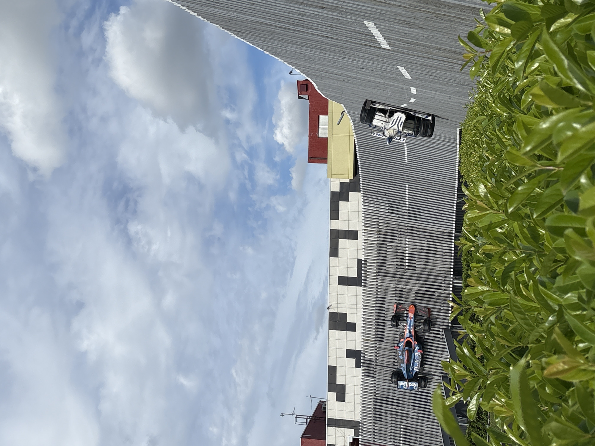 A high-angle shot depicts a miniature racetrack setup outdoors. Two Formula 1 cars are visible: one is predominantly red and blue, and the other is white and black. The track runs parallel to a building with a checkered pattern on its side. The sky is visible above the building and is filled with white clouds. Green foliage lines the side of the track. The location is Anapa, Russia.