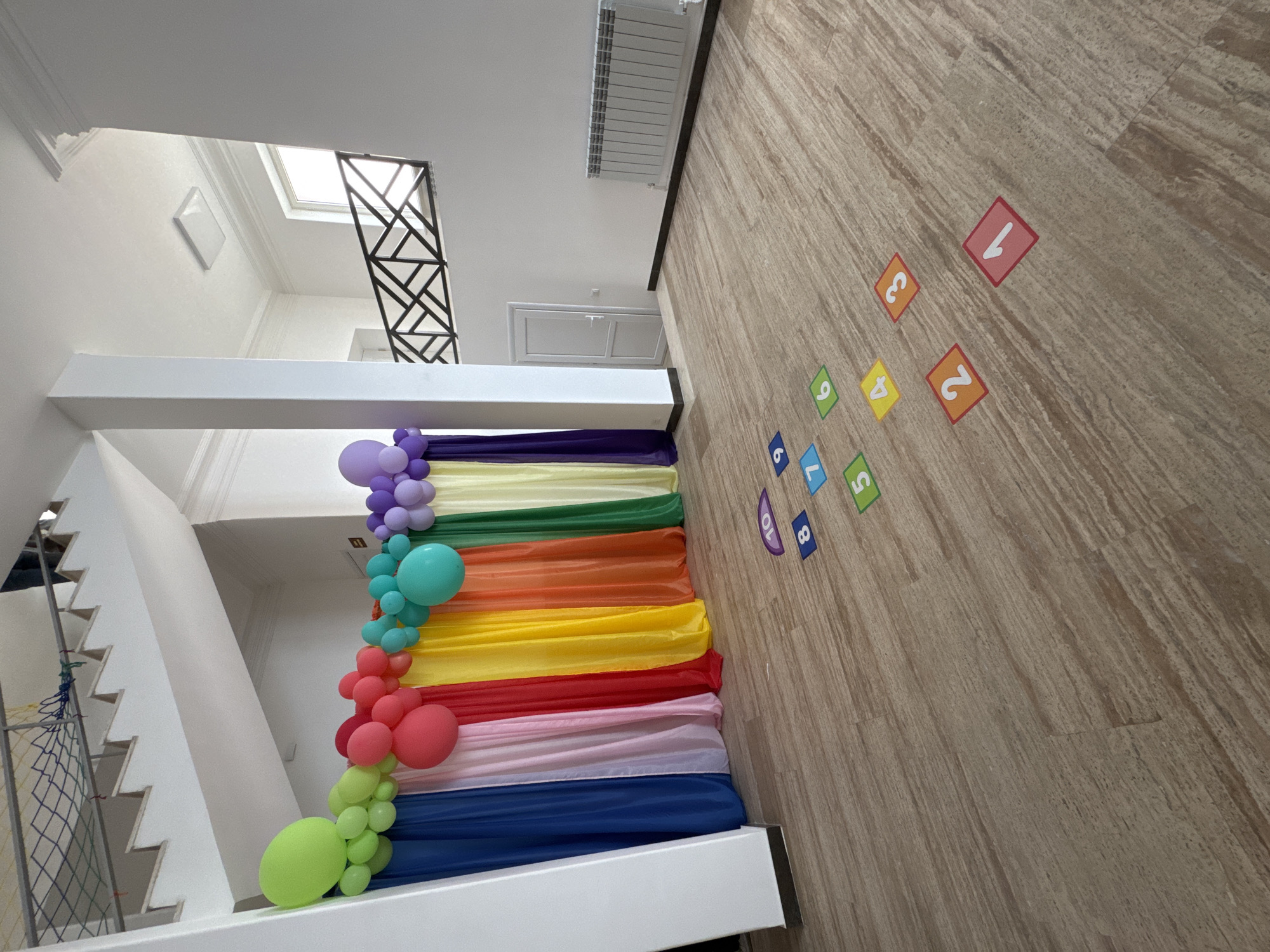 The image displays a brightly decorated interior space, possibly a hallway or staircase landing in a multi-level building located in Yerevan, Armenia. The primary focus is a vibrant rainbow-themed decoration consisting of draped fabric panels and a corresponding balloon garland. The fabric starts with deep blue at the bottom, progressing through light pink, red, yellow, orange, green, light yellow, lavender, and culminating in dark purple. The balloon garland features clusters of balloons matching these colors, creating a festive arch.
To the left, a white staircase with a modern black geometric metal railing ascends. The walls and ceilings are white, and natural light appears to enter through a skylight visible through a ceiling opening, suggesting it is daytime. A white radiator is mounted high on a wall, and a white ceiling hatch is also present.
The floor is covered with light brown, wood-patterned tiles or laminate. Scattered across the floor are colorful square decals featuring numbers 1 through 10. The numbers are presented on different colored backgrounds: red (1, 3), orange (2, 5), yellow (4, 7), green (6, 9), and blue (8, 10). These floor markings suggest an area designed for children's play or learning activities.
The overall atmosphere is cheerful and celebratory, indicating the space is prepared for an event such as a birthday party, children's gathering, or a dedicated play area. No people are visible in the scene.