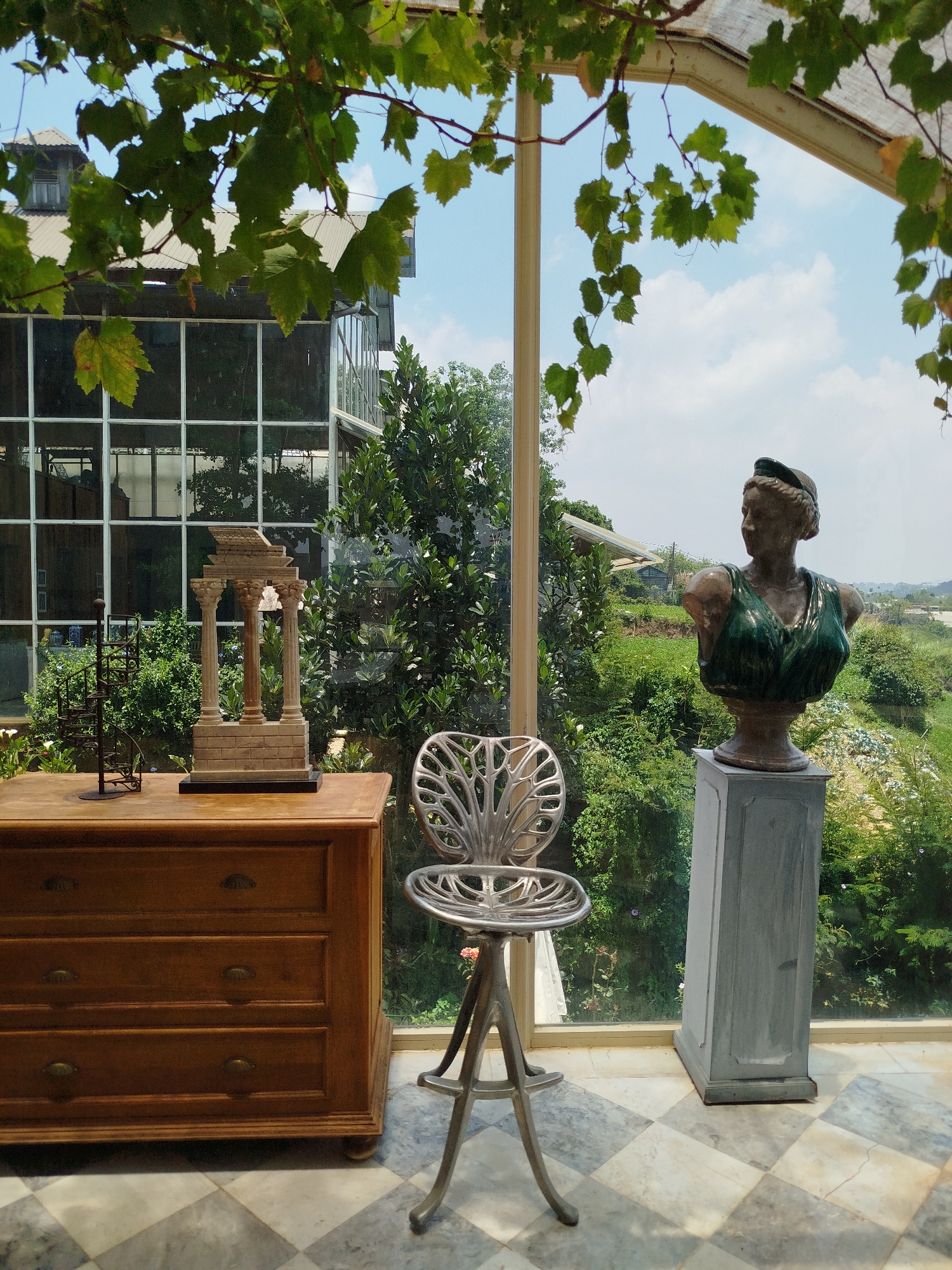The image presents an interior space, likely a conservatory or sunroom, characterized by large glass windows that offer an expansive view of a verdant outdoor landscape, typical of Lang Biang, Đà Lạt, Vietnam.
In the foreground, several decorative objects are arranged. A wooden dresser with three drawers and brass handles stands on the left. It supports a miniature architectural model resembling a classical temple with columns, alongside a dark, spiral metal sculpture. A distinctive metallic chair or stool, featuring an ornate, leaf-patterned seat and three legs, occupies the center. To the right, a bust of a woman, appearing to be made of bronze or terracotta with green drapery, is displayed on a tall, grey pedestal. The floor throughout the indoor area is laid with a checkered pattern of light and dark grey tiles. Green vines with numerous leaves hang from an overhead structure, partially obscuring the sky and casting natural shadows.
Beyond the glass, a lush, hilly terrain unfolds under a bright, partly cloudy sky, indicating daytime. Dense green foliage and trees are prominent just outside the windows, with further hills covered in vegetation visible in the distance. A building with a corrugated roof and grid-patterned windows is discernible through the leaves in the upper left background.
No people or visible text are present in the image. The scene exudes a serene, sunlit atmosphere that harmoniously blends indoor decor with the natural beauty of the Vietnamese landscape.