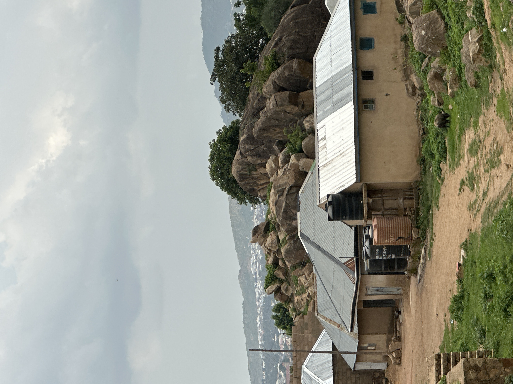 This image captures a landscape in Jos, Nigeria, featuring buildings nestled against a rocky, vegetated hillside under an overcast sky. The photograph is oriented vertically, showing a large portion of the sky to the left and the ground with structures to the right.
In the foreground and midground, several buildings with light-colored, possibly sand-colored, walls and shiny corrugated metal roofs are visible. The most prominent building on the right has multiple small, dark-framed windows. Adjacent to or on top of these buildings are several water storage tanks, including a rusty-brown cylindrical tank and two black tanks. On one of the black tanks, faint white text, possibly indicating capacity like "500 L" or "Water Tank," can be partially discerned. A utility pole extends upwards from near the lower center.
Behind the structures, a rugged hillside rises, covered in large, rounded boulders and sparse green vegetation, including bushes and grass. A dark-colored animal, possibly a goat or small livestock, is seen grazing on a patch of grass near the building on the right.
In the background, beyond the immediate hills, a sprawling settlement of buildings is visible in a valley or on distant slopes, suggesting a populated area. The sky is a uniform light grey, heavily overcast with some brighter cloud patches, indicating a cloudy daytime, possibly late morning or afternoon, with soft, diffused light. There are no people visible, and no specific activities other than the animal grazing are taking place.