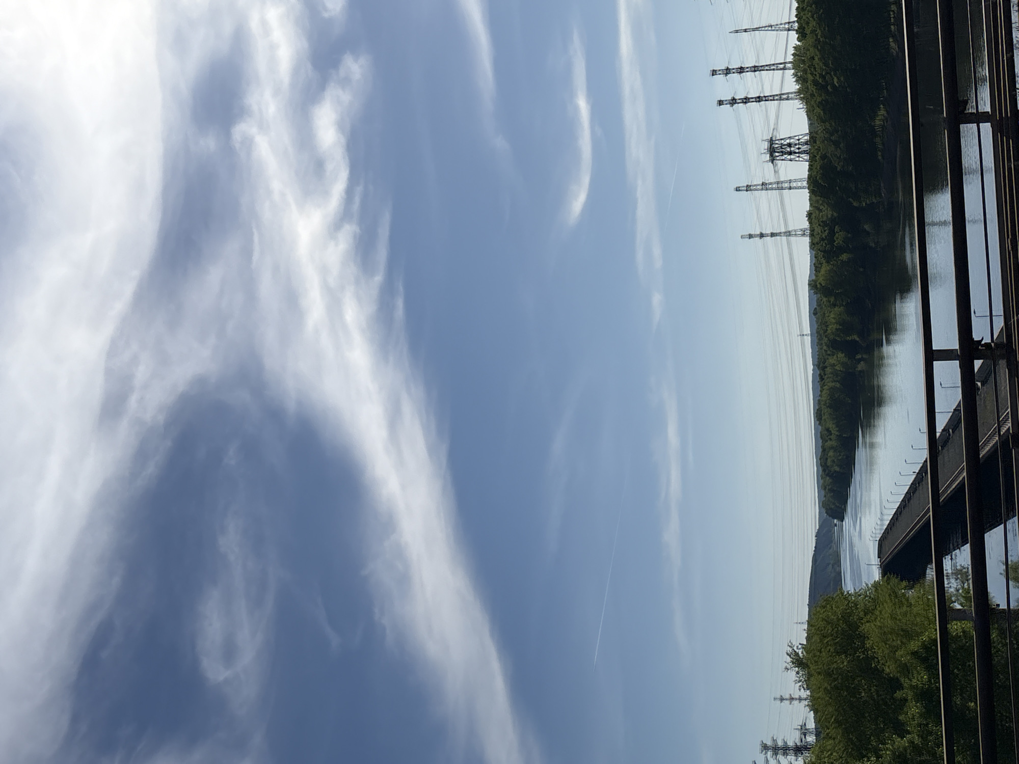 The image shows a sky with cirrus clouds. A line, perhaps a contrail from an airplane, is visible against the blue sky. In the background, there is a body of water, possibly a river or large lake. The presence of power lines and pylons suggests this is a landscape with some level of industrial development. A tree line borders the water. Vertical bars are present in the right of the image. The location is likely Chaikovsky, Russia due to the presence of power lines.