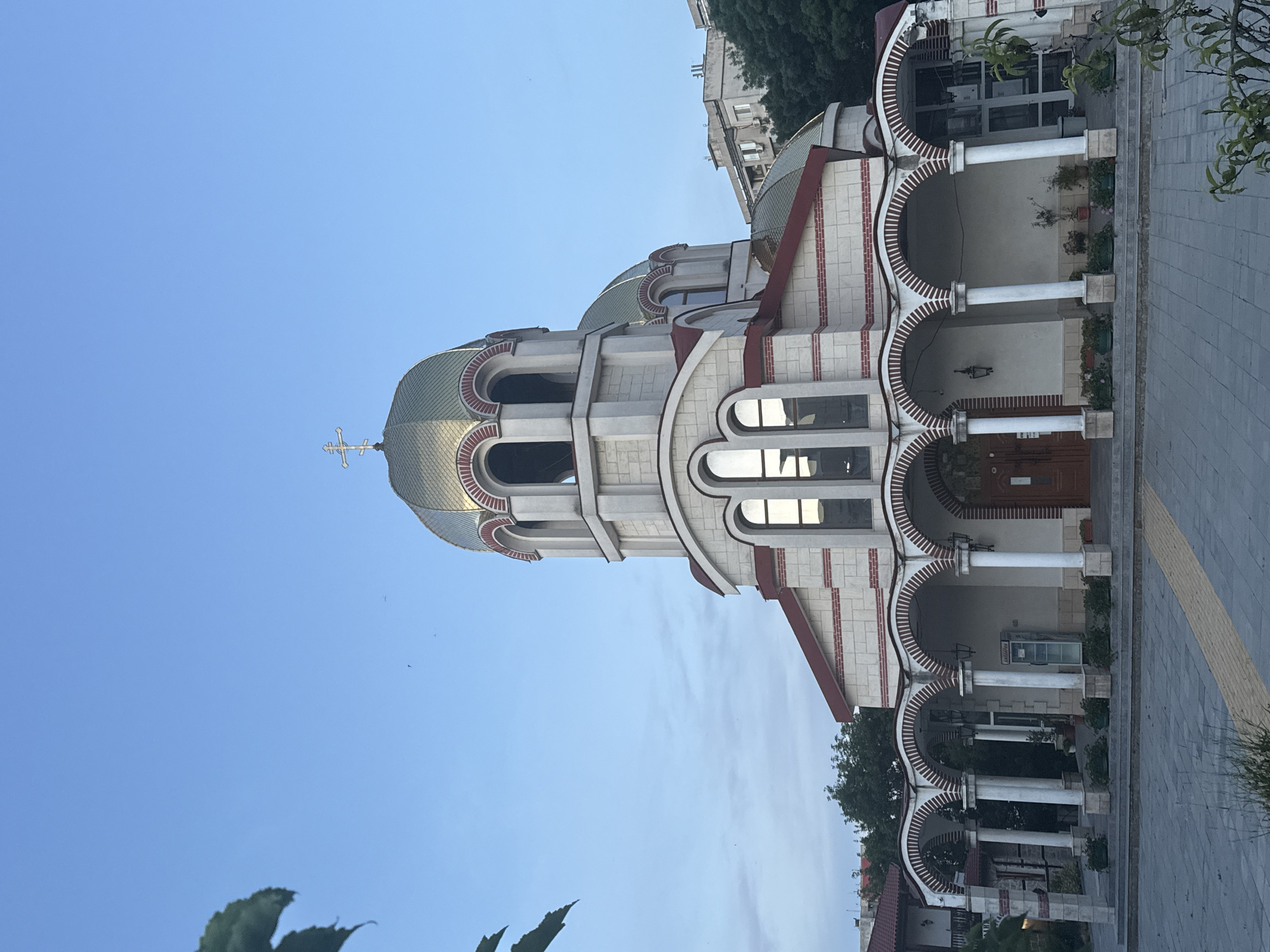 The image shows the exterior of the Saint Nicholas the Miracleaker Russian Orthodox Church located in Burgas, Bulgaria. The church features a prominent golden dome topped with a cross, and a facade constructed with light-colored stone accented by red brickwork. An arched colonnade with white pillars runs along the front of the building, providing a covered walkway. The church is situated in an urban setting, with trees and other buildings visible in the background under a clear blue sky. The scene is devoid of any people.