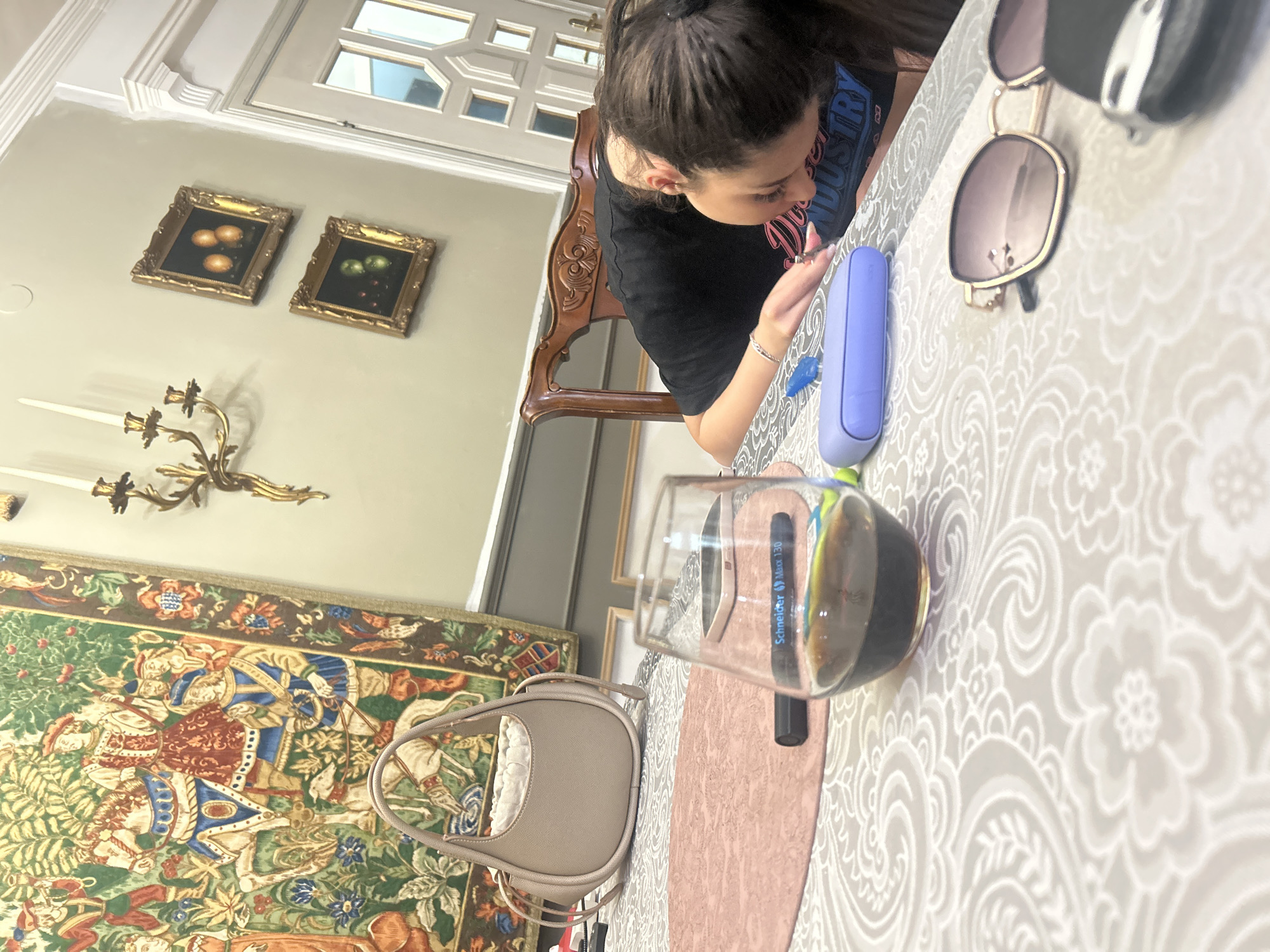 The scene depicts an interior setting, specifically a dining or living room within a residence or establishment in Plovdiv, Bulgaria. A single female subject, identifiable by dark hair tied in a ponytail, is seated at a table, leaning forward intently over an object directly in front of her, the object itself obscured by her body and focus. She is wearing a black t-shirt with partial lettering "DUSTRY" visible in blue and pink. A silver-colored bracelet is present on her right wrist.
The table is covered with a white tablecloth featuring a subtle, intricate pattern of floral and swirling motifs. A pink, oval-shaped placemat is positioned beneath several items. On the table, from foreground to background, are: a pair of gold-framed sunglasses with dark lenses, a black car key fob, and a light purple/blue elongated case. To the left of these items, a clear glass partially filled with a dark liquid, consistent with red wine or a dark beverage, is prominently displayed. Behind the glass, a black marker with "Schneider Maxx 130" printed on it rests on the placemat. Further back on the table, a light beige/taupe handbag with a drawstring closure is visible.
The background wall features a large, ornate tapestry depicting a vibrant historical or mythological scene with multiple figures, including individuals on horseback, pedestrians, and a dog, set against a verdant landscape. To the right of the tapestry, a gold-colored wall sconce with three white candles is affixed to the wall. Below the sconce, two framed still-life paintings hang vertically: the upper painting displays three orange spherical fruits, and the lower one depicts green apples and red cherries, both within ornate gold frames. To the far right, a white paneled door with glass inserts is visible, indicating passage to another room. The overall environment presents a classical and detailed aesthetic.
