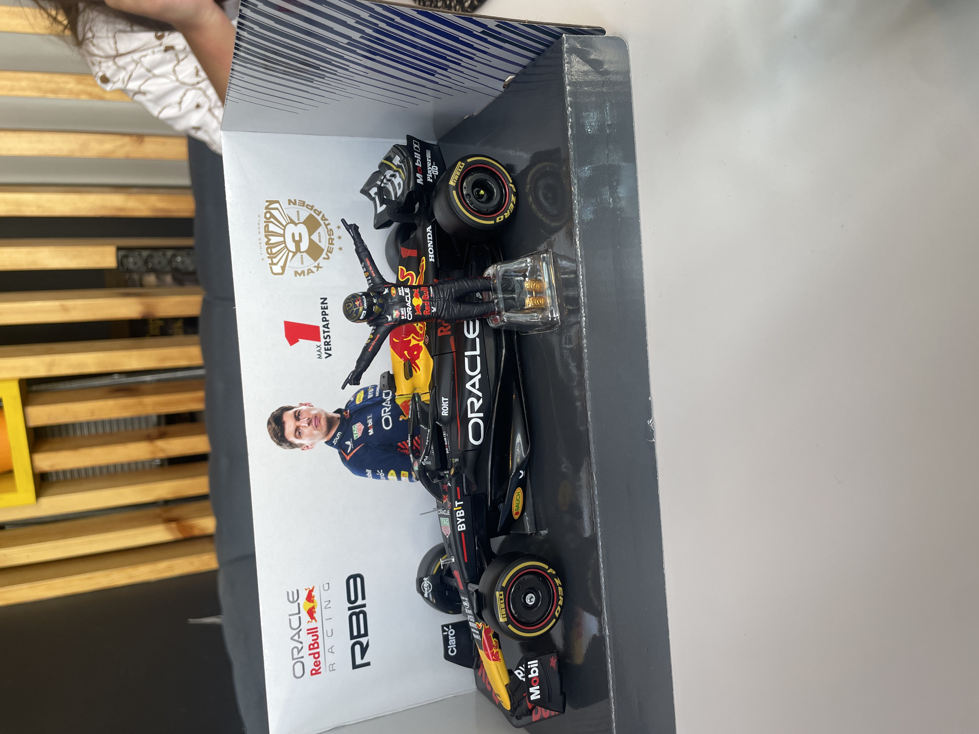 A miniature display of a Formula 1 car and driver is shown on a table. The display is housed within a box featuring the Oracle Red Bull Racing logo, the RB19 car model, and a picture of Max Verstappen. A small figurine of Max Verstappen in his racing suit, with arms outstretched, stands in front of the car model. The car itself is black with red and yellow accents, and features various sponsor logos. The background includes a blurred image of wooden slats and part of a person wearing a patterned shirt. The scene is lit by overhead lighting.