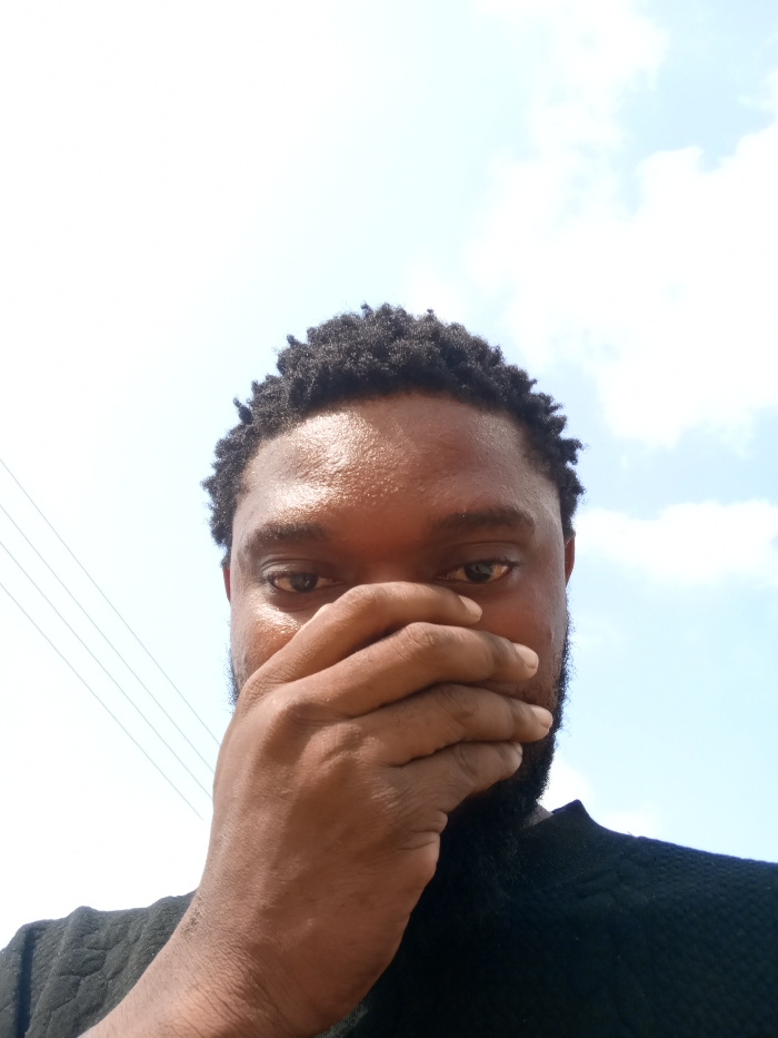 An adult male subject is depicted in a close-up, upward-angled outdoor portrait taken in Magboro, Nigeria. He has dark skin, short, kinky dark hair, and a dark beard. His right hand is positioned over his lower face, obscuring his mouth and nose. His dark eyes are open and gaze directly forward. He is wearing a dark, textured garment. The background consists primarily of a bright, clear sky, with scattered white clouds present on the right side. Several dark, thin utility wires are visible in the upper-left portion of the frame.