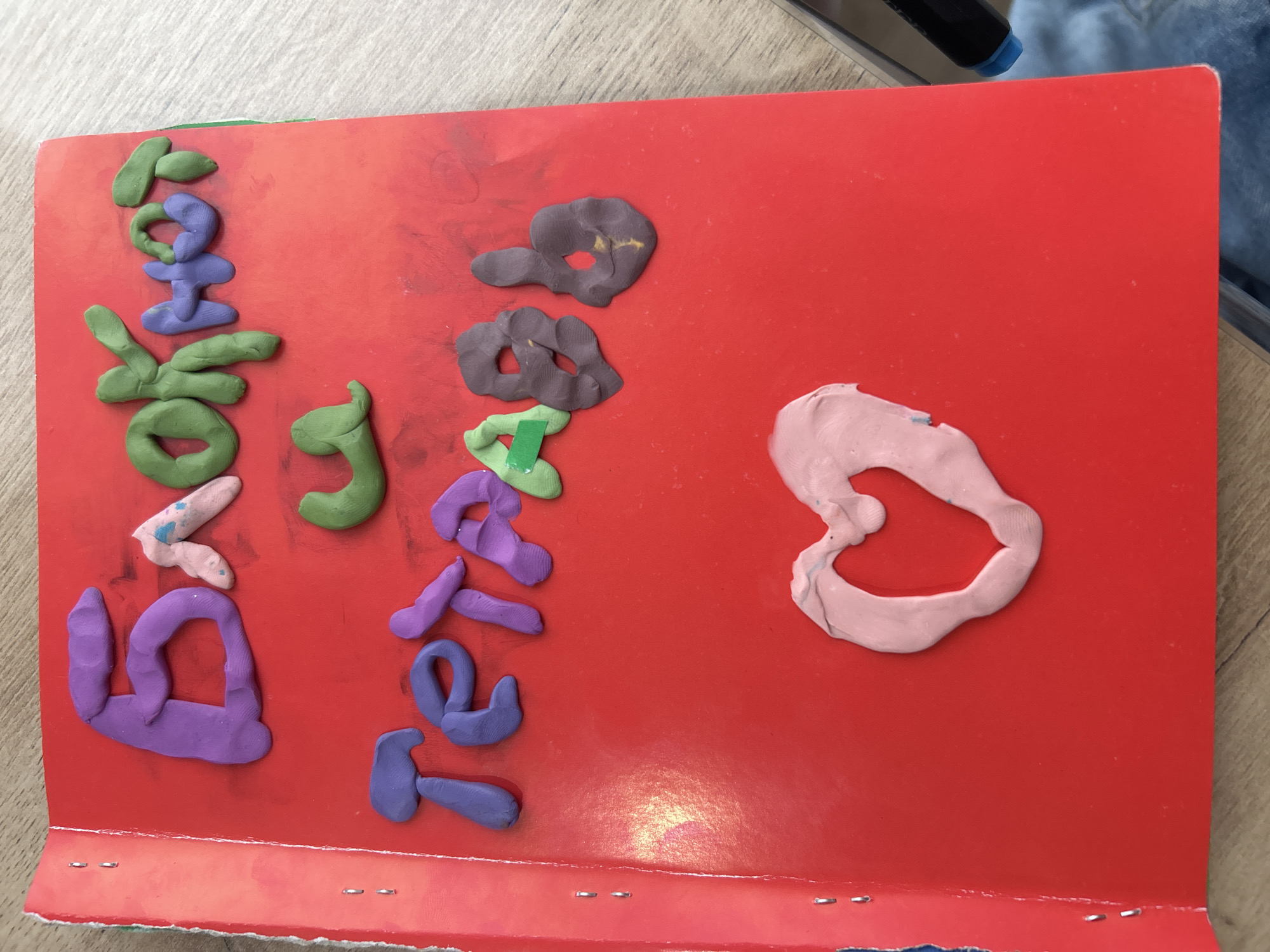 A red, stapled-edge notebook sits on a wooden surface. The notebook cover is decorated with colorful modeling clay shaped into what appears to be letters and shapes. From left to right, vertically, the first word appears to be "Doklad" (Доклад in Russian), the second looks like "Tezis" (Тезис in Russian), and a heart shape is situated towards the right side of the notebook. No people are visible. The notebook is placed diagonally with the bottom left corner closest to the viewer.
The clay letters are of various colors including purple, green, light pink, and brown. The heart is in pink. The notebook's background is a bright red color. No city or specific location can be identified in the provided image.