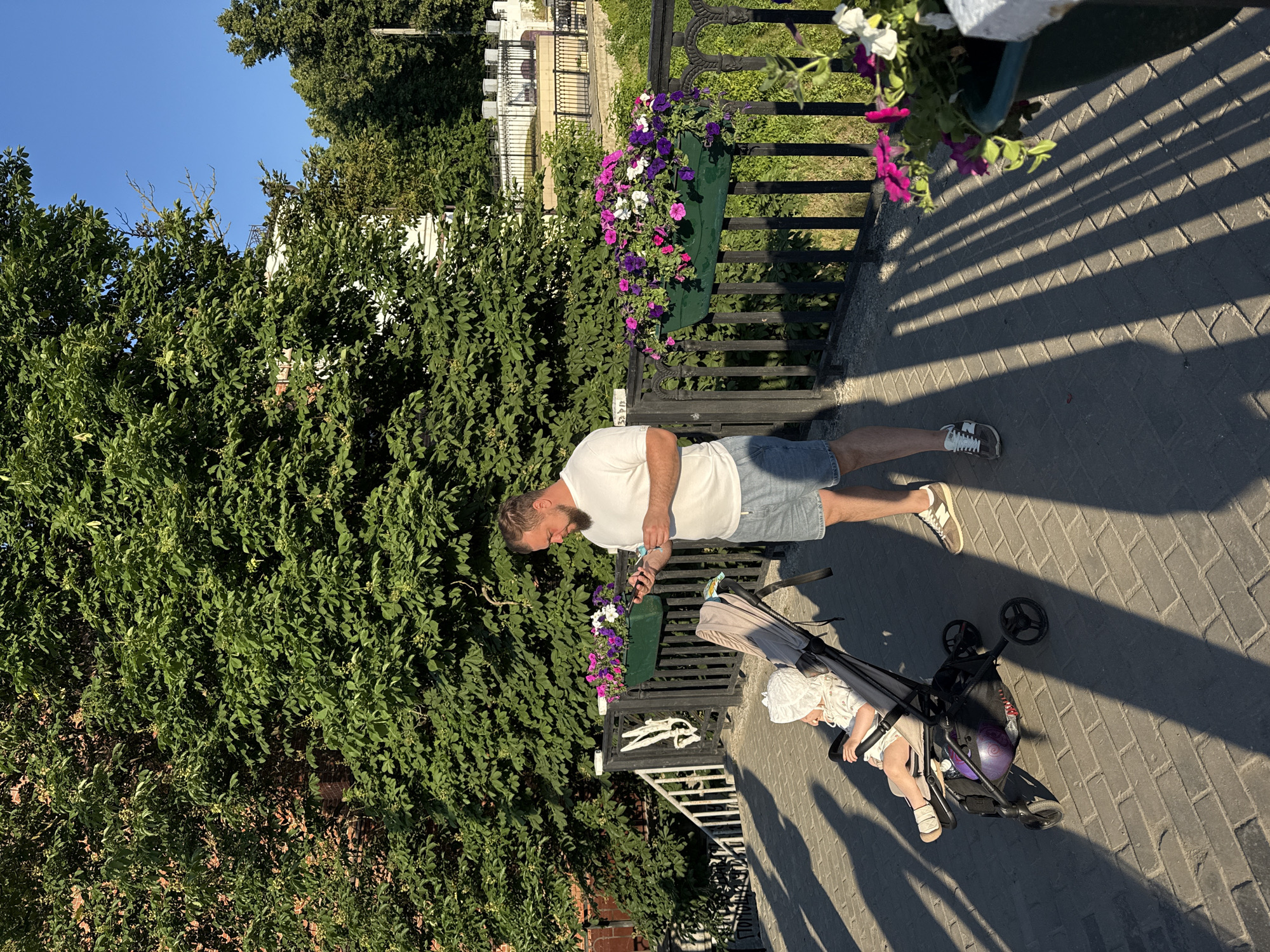 The image depicts a man standing on a paved walkway, holding a mobile phone, next to a stroller containing a young child. The man is wearing a white t-shirt and denim shorts. The child is dressed in light-colored clothing and a hat. In the background, a black metal railing with flower boxes filled with colorful petunias is visible. Behind the railing, there are stairs leading down and lush green trees. The sun casts long shadows on the walkway, indicating it is late afternoon or early evening. The architecture in the distance suggests an urban setting, possibly a park or a promenade. The inscription "ПОМНИМ" (which translates to "We Remember") is visible on a staircase to the left of the frame. This detail, combined with the overall ambiance, suggests the location might be a memorial or a significant public space within the city of Vladimir, Russia.