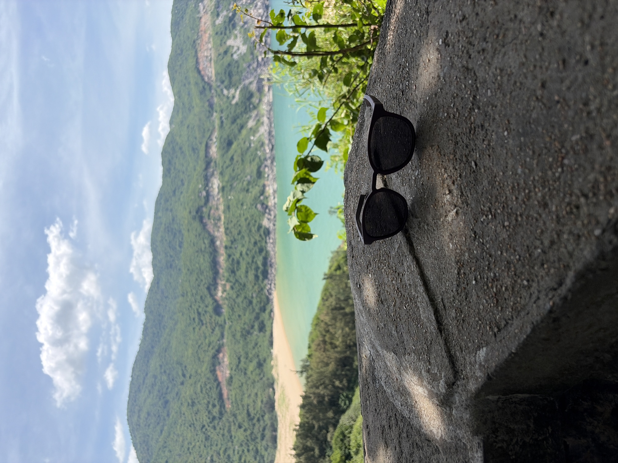 A pair of black sunglasses rests on a textured, grey stone surface in the foreground. Behind the sunglasses, a scenic view unfolds with a turquoise ocean, a light-colored sandy beach, and lush green mountains under a cloudy blue sky. The location is identified as Hòa Xuân, Vietnam. The image is taken from a slightly elevated perspective, with the sunglasses positioned on what appears to be a ledge or wall overlooking the coastline.