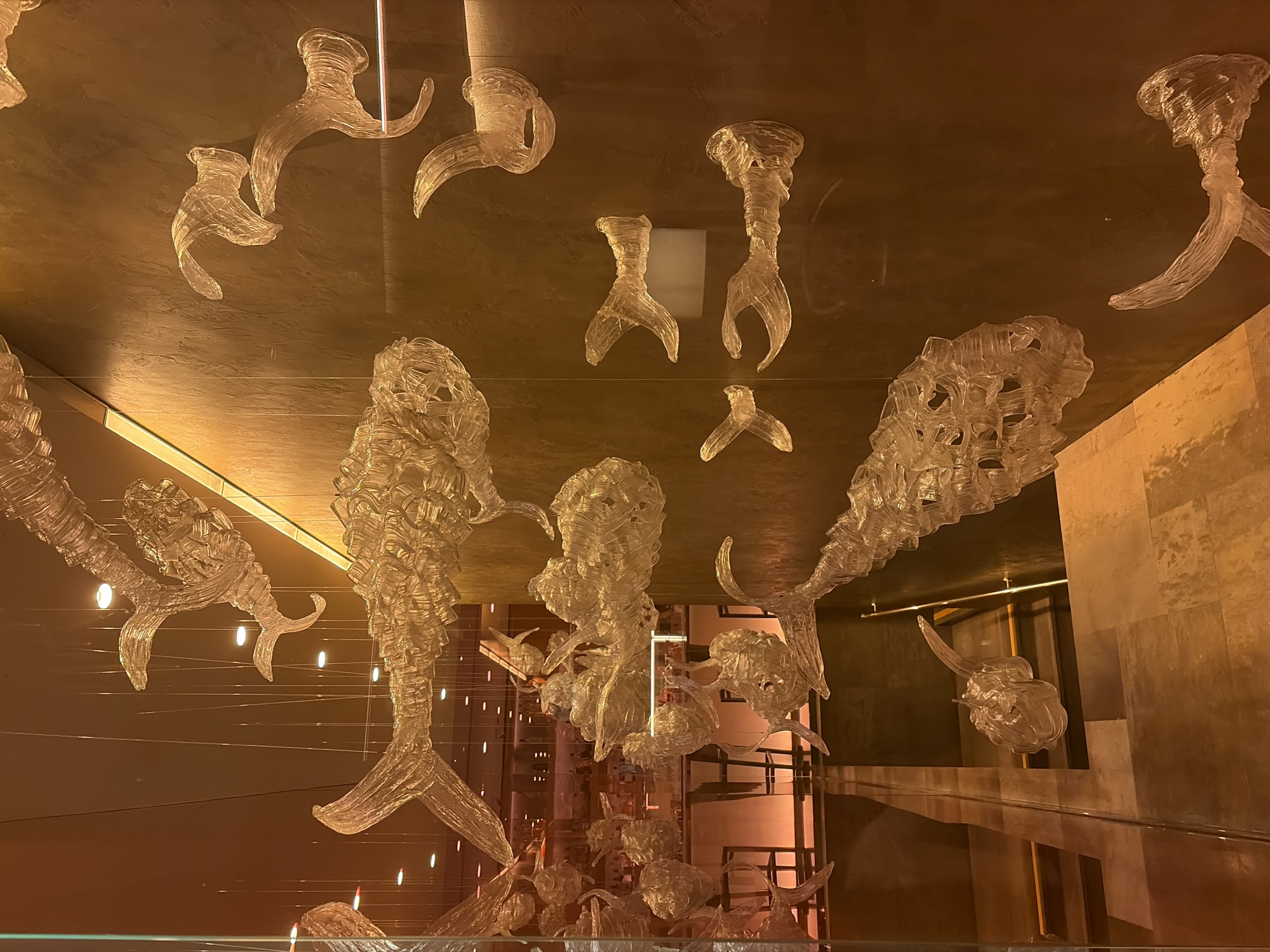 The image displays a wall art installation of transparent, sculpted glass fish, suspended at various heights. The fish are crafted in a detailed, segmented style, giving them a shimmering, almost skeletal appearance. The background is a warm, textured wall with ambient lighting, reflecting the glass sculptures. In the background, visible through what appears to be a glass partition, is a bar area with stools and bottles, illuminated with soft, warm lights. This scene is located at the bar at the Four Seasons Hotel Lion Palace in Saint Petersburg, Russia. There are no people visible in the immediate foreground of the image.