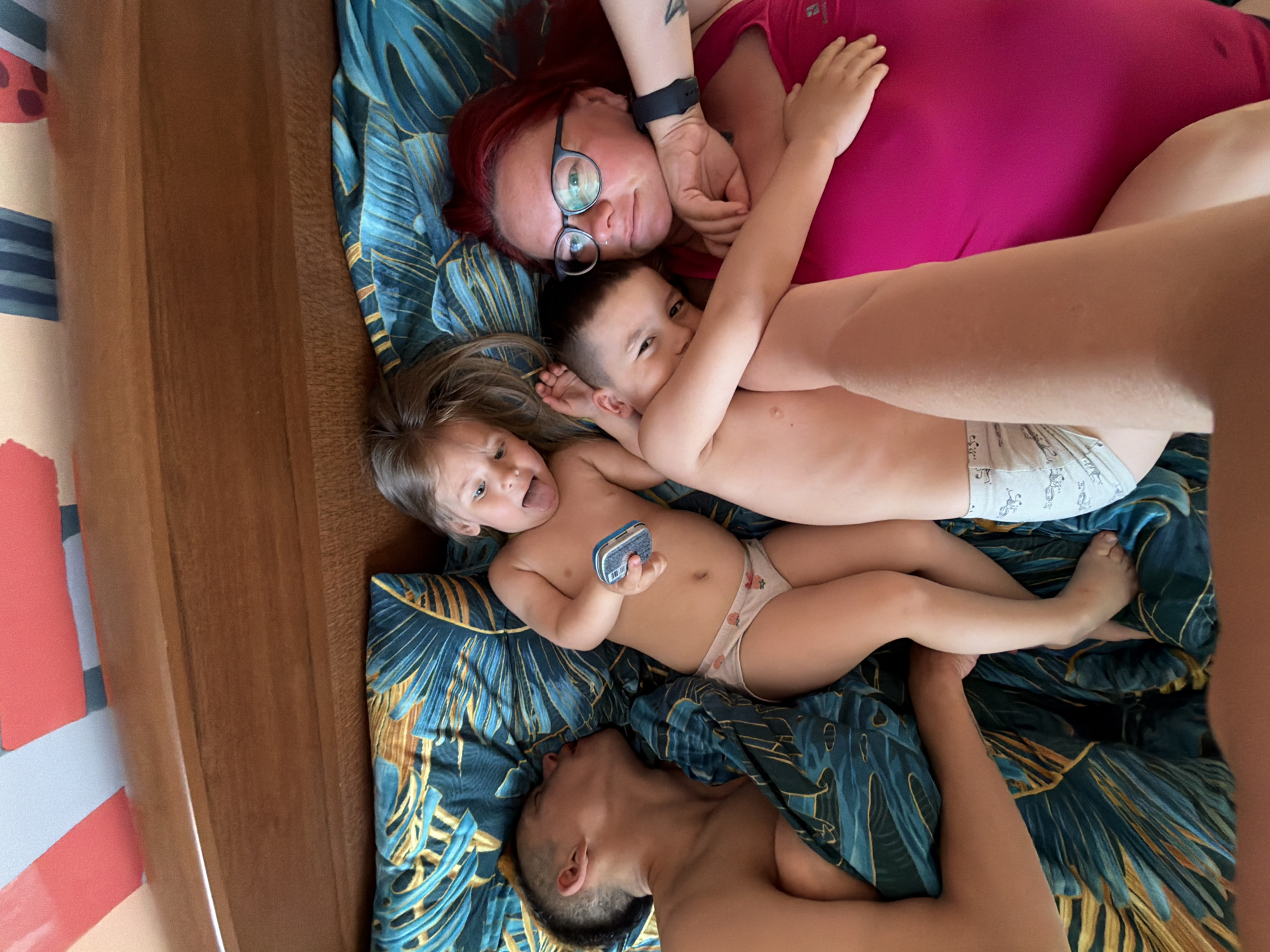 A woman with red hair and glasses in a pink shirt is lying on a bed with a patterned blue and gold duvet, embracing two young children. One child, a girl with blonde hair, is sticking her tongue out and holding a small rectangular object. The other child, a boy, is looking at the camera. A man is also lying on the bed. The location is Chaikovsky, Russia.
