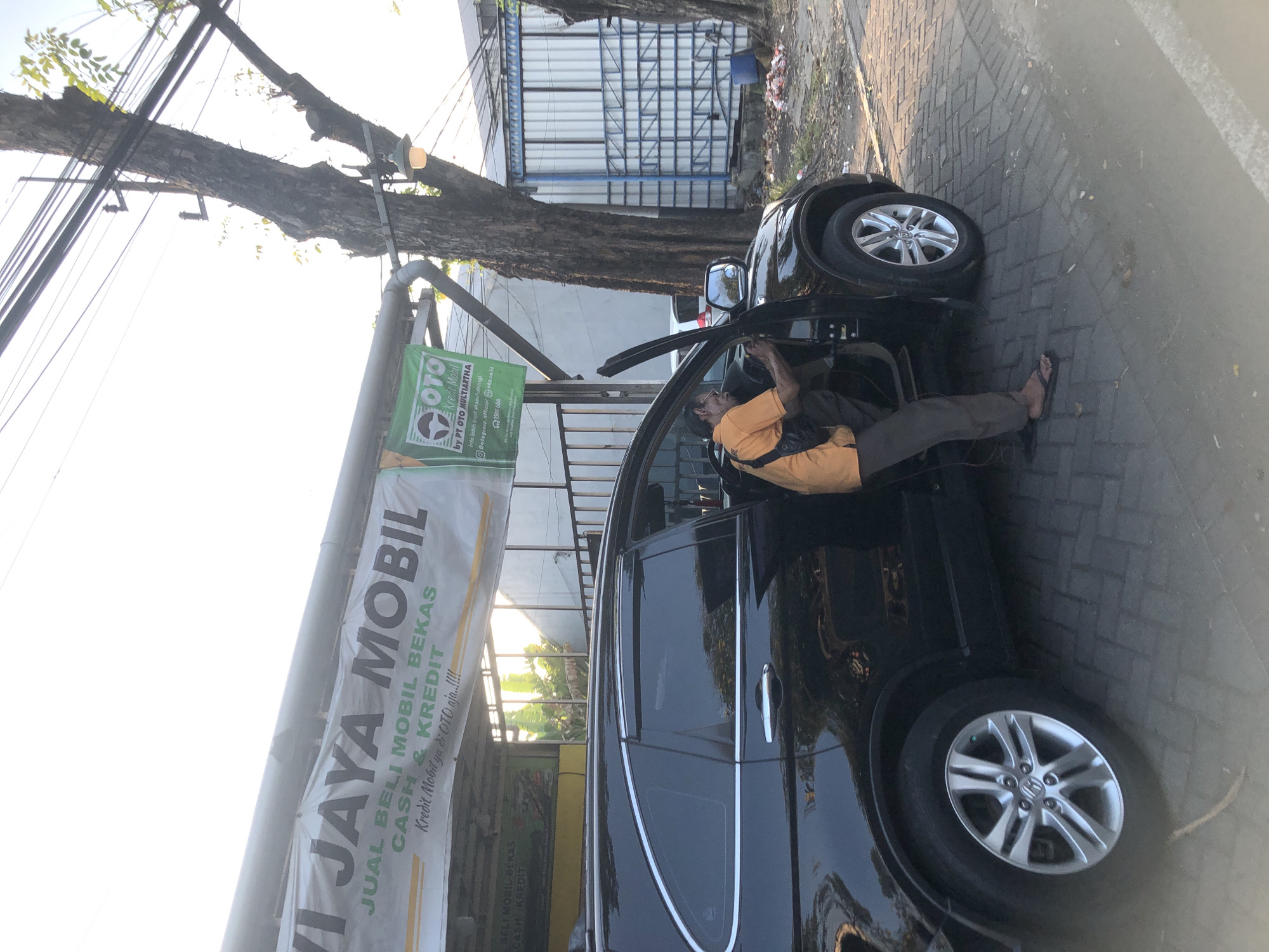 A man in an orange shirt and gray pants is working on the driver's side of a black SUV. The SUV has its door open and the man is reaching into the interior. He is wearing flip-flops. The car is parked on a brick-paved surface. Above the car is a banner that reads "JAYA MOBIL JUAL BELI MOBIL BEKAS CASH & KREDIT". To the right of the banner is a green sign with the OTO logo and text that says "Kredit Mobil via OTO aja....!!!" and "by PT OTO MULTIARTHA". In the background, there are trees, electrical wires, and a building with a blue and white corrugated metal facade. The scene appears to be outdoors, possibly at a car dealership or service center, given the banner and the man's actions. The lighting suggests it is daytime, likely late afternoon due to the warm glow and long shadows. The location is identified as Gelam, Indonesia.