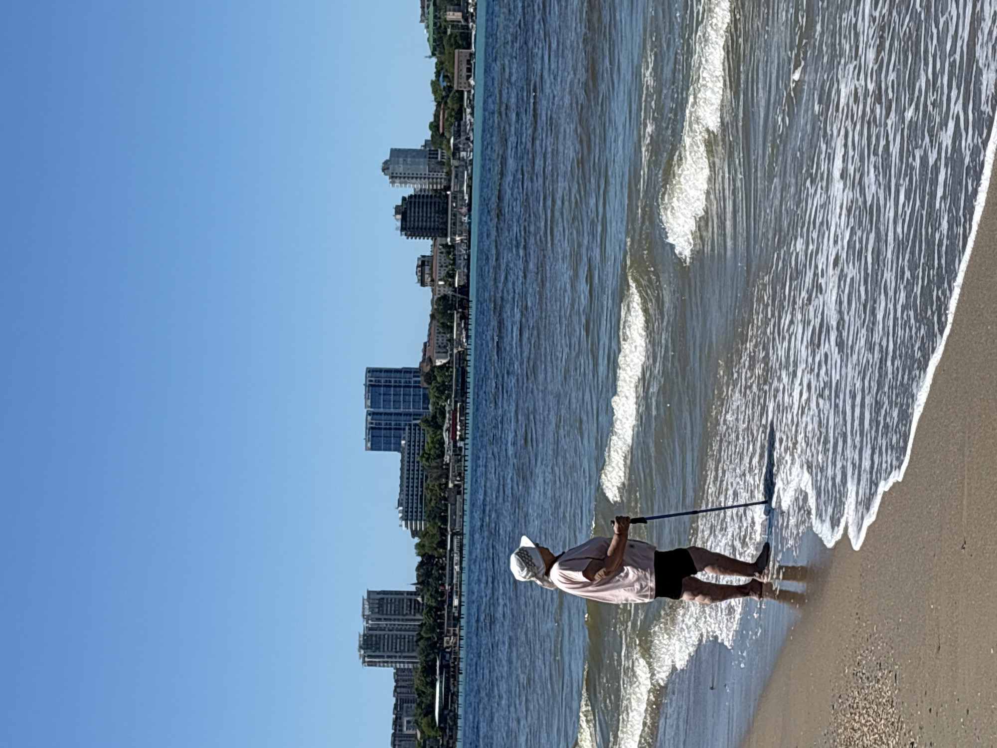 An image captured in Anapa, Russia, shows a person standing at the edge of the Black Sea, where gentle waves meet a sandy beach. The image is rotated 90 degrees clockwise.
The main subject is an adult, clad in a light-colored short-sleeved shirt and dark shorts, with a white or light-colored cap on their head. They are barefoot, with their feet partially submerged in the shallow water as small waves break around them. The person is holding a long, dark pole or stick, which extends into the water ahead of them.
To the left, beyond the immediate shoreline, the sea stretches out under a clear, bright blue sky. In the background, along the distant coastline, a modern cityscape is visible, characterized by several multi-story buildings and what appears to be a developed urban waterfront. The weather is sunny and clear, indicative of a pleasant day, likely morning or afternoon, given the bright light and lack of long shadows. No discernible text is visible within the image. The scene conveys a sense of leisure and coastal activity.