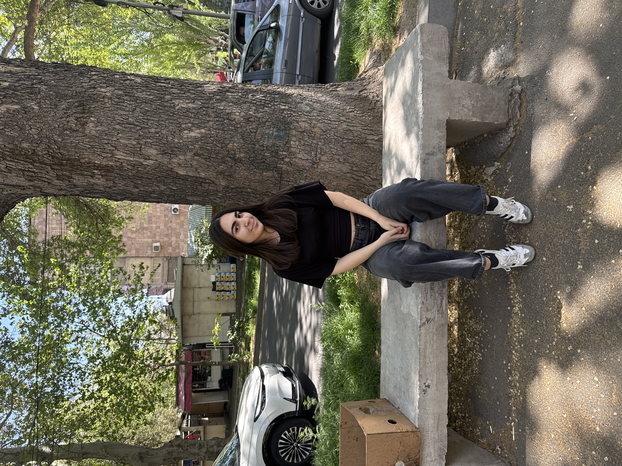 A woman is sitting on a concrete bench next to a large tree in what appears to be Yerevan, Armenia. She is wearing a black top, grey jeans, and white sneakers. She is looking at the camera with a slight smile. Behind her, there is a street with cars parked along the side. Buildings can be seen in the background. A cardboard box is located next to the bench. The ground is paved and has spots of sunlight.